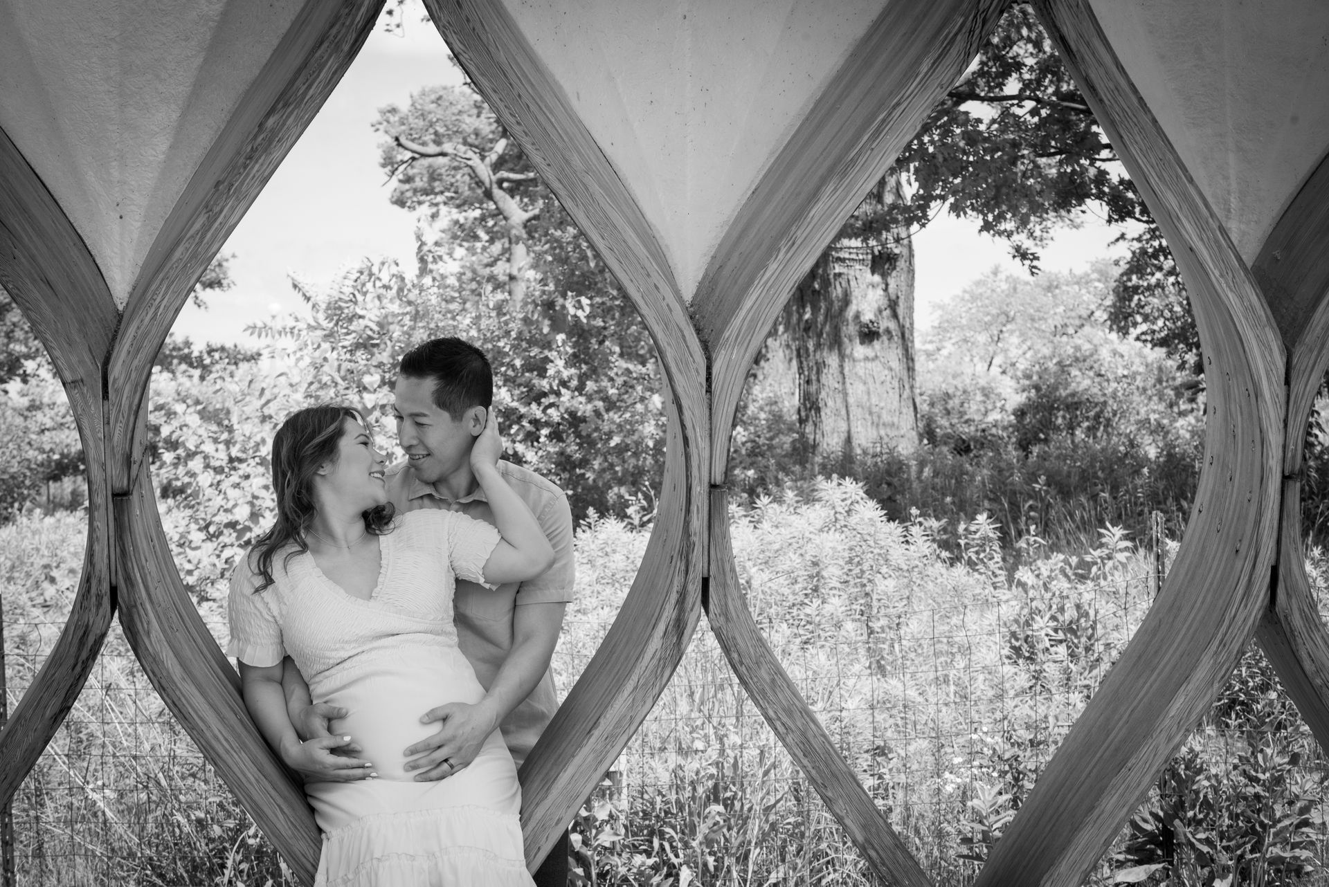 Pregnant couple embracing, framed by a wooden structure with a nature background.