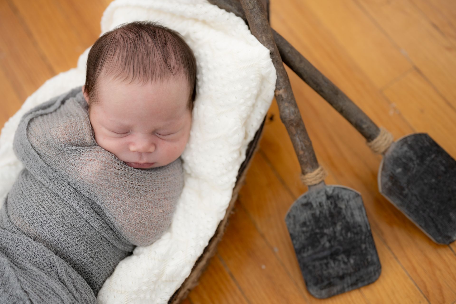 Newborn swaddled in gray blanket, resting in a wooden bowl with two oars on a wood floor.