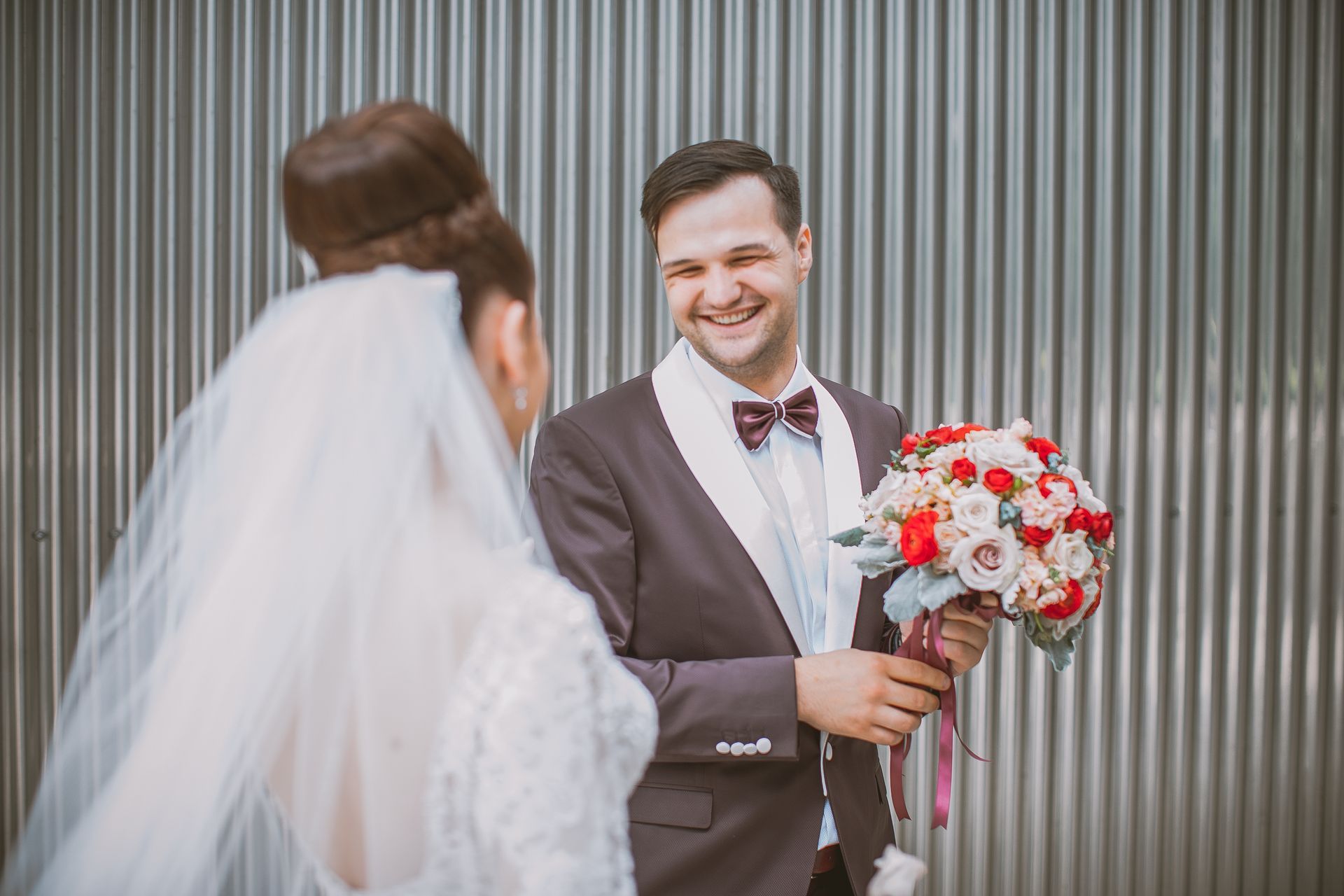 Groom smiles, holding flowers, facing bride in veil. They stand before a corrugated metal wall.
