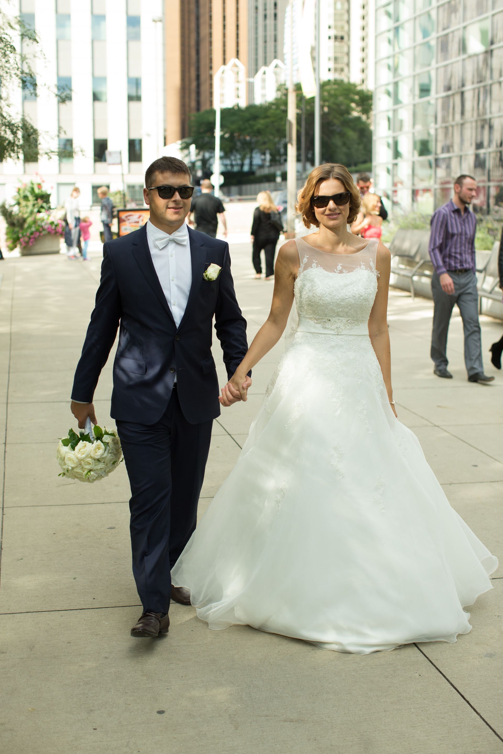 Newlyweds walk hand-in-hand outdoors. Groom in suit holds flowers. Bride wears a white wedding dress. Sunny day.