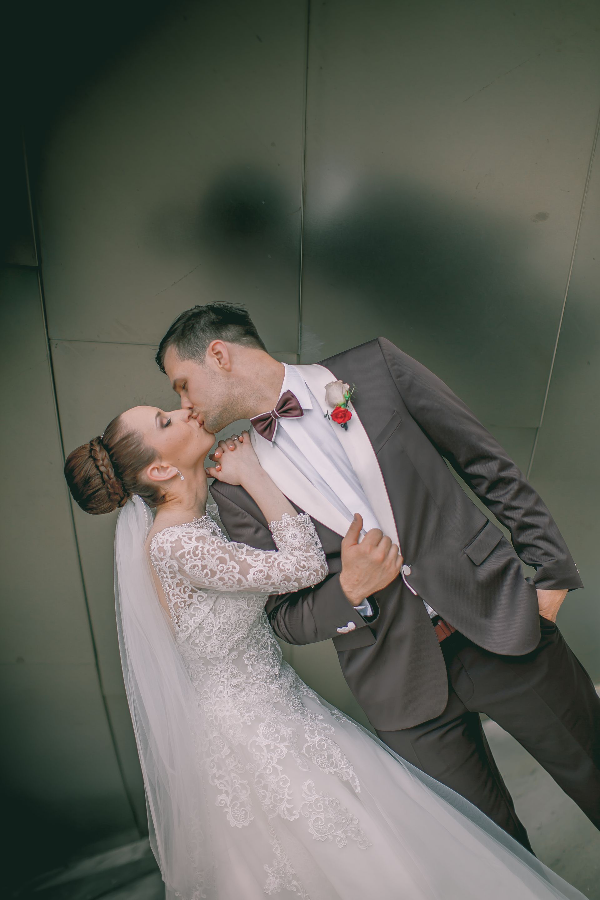 Wedding couple kissing in front of a gray wall. The bride wears a white dress and veil, the groom a brown suit.