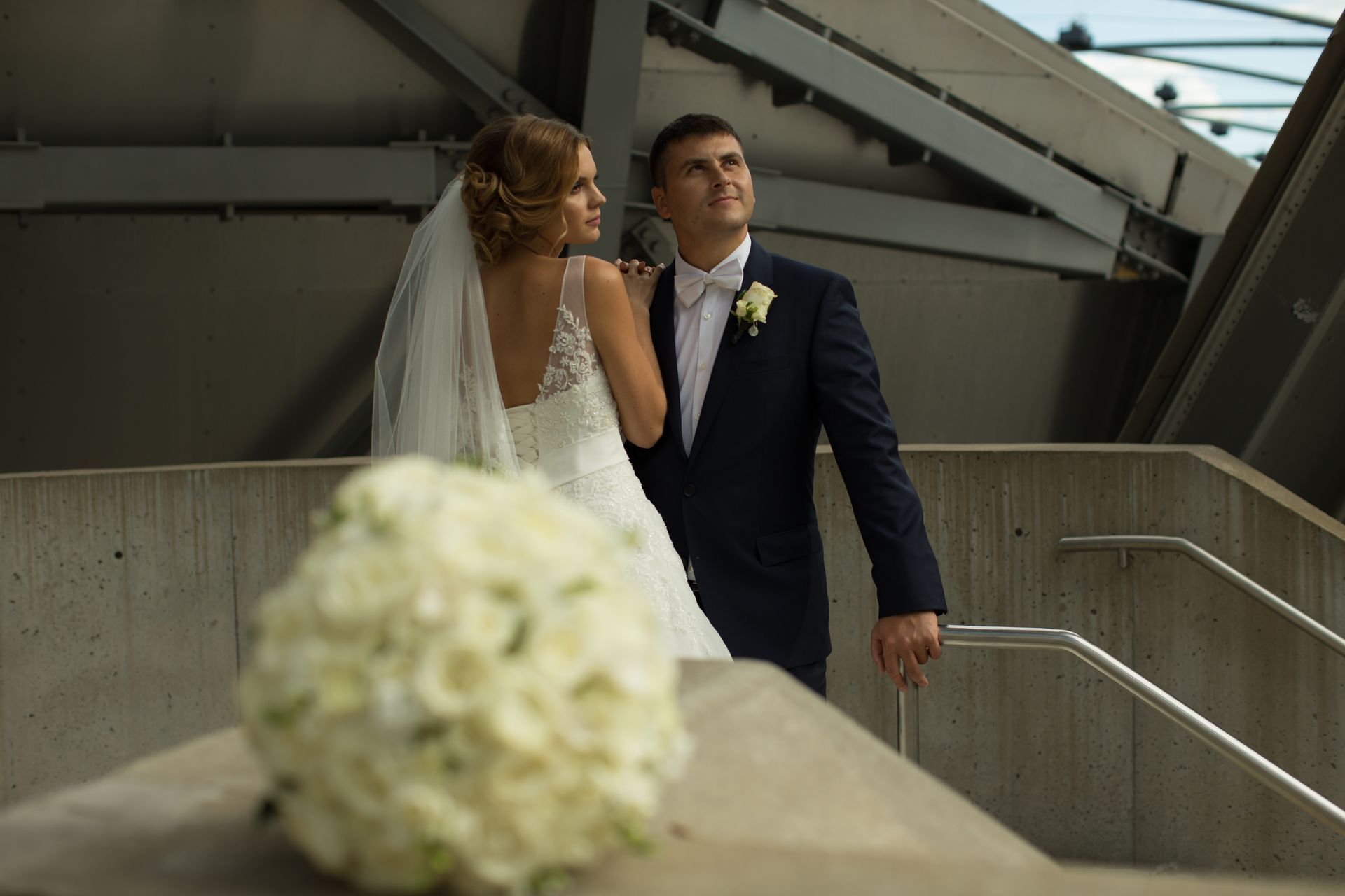 Wedding couple on concrete steps; bride in white dress, groom in suit, looking up.