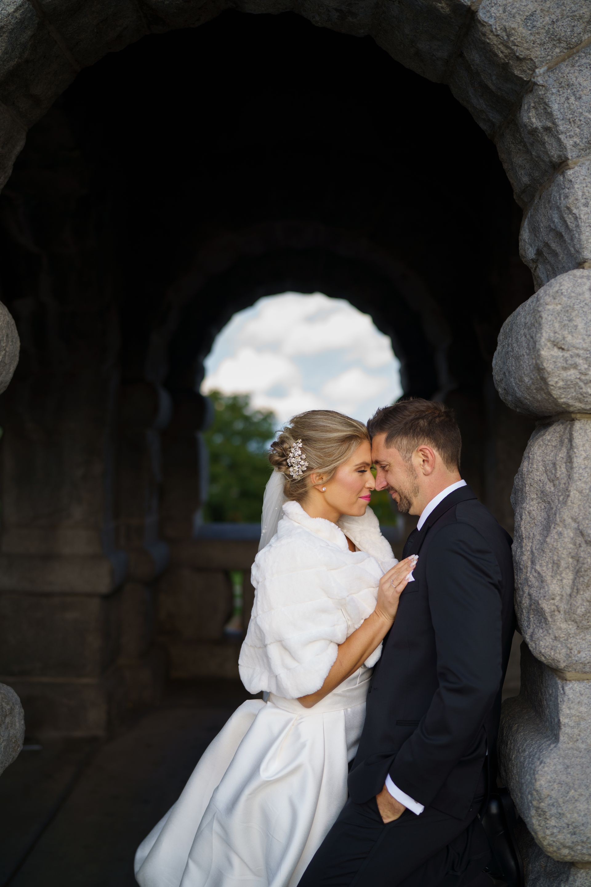 Bride and groom embracing in stone archway, soft white dress, dark suit, intimate moment.