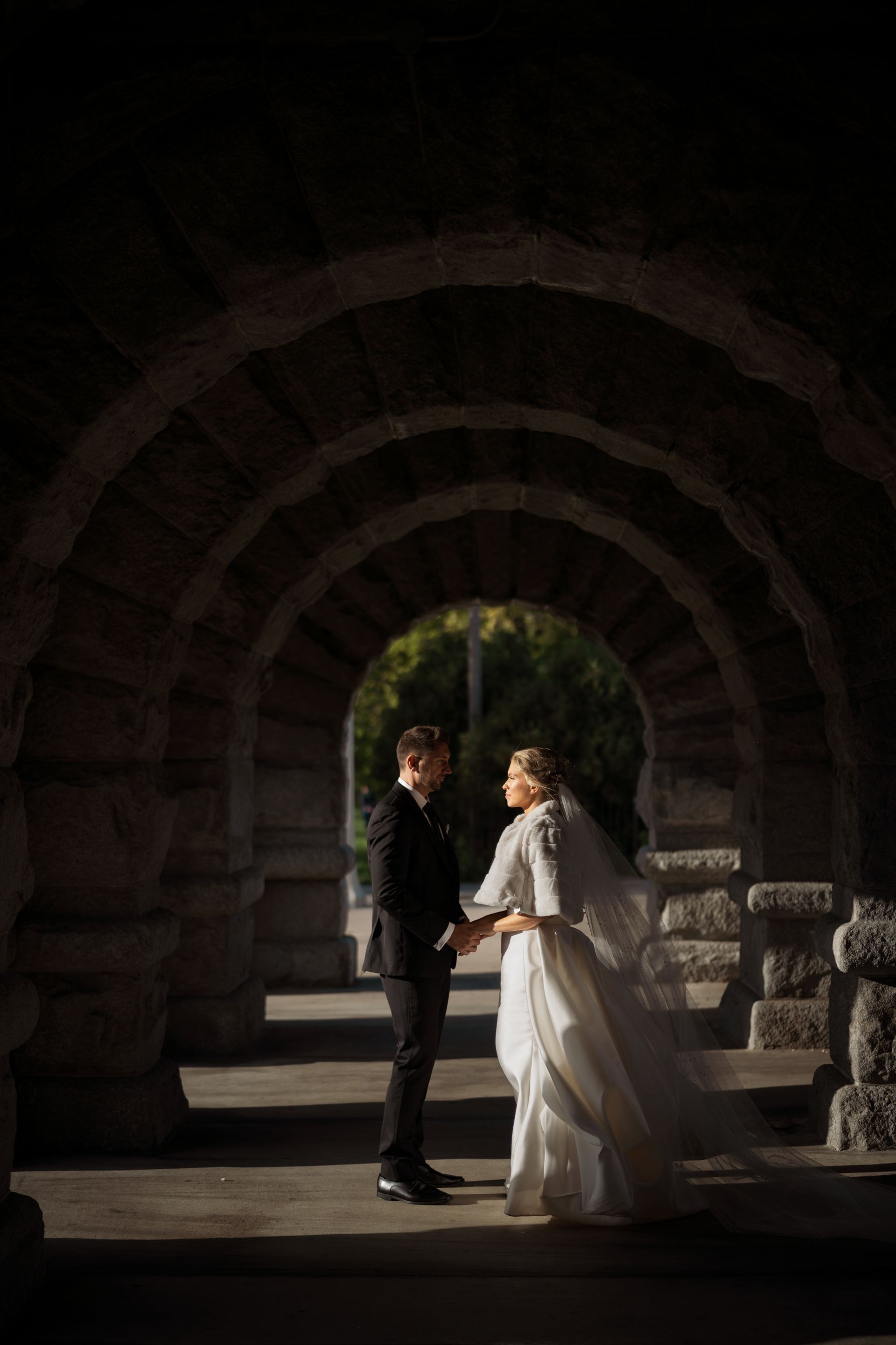Couple holding hands under stone arches; bride in white gown, groom in black suit. Sunny daylight.