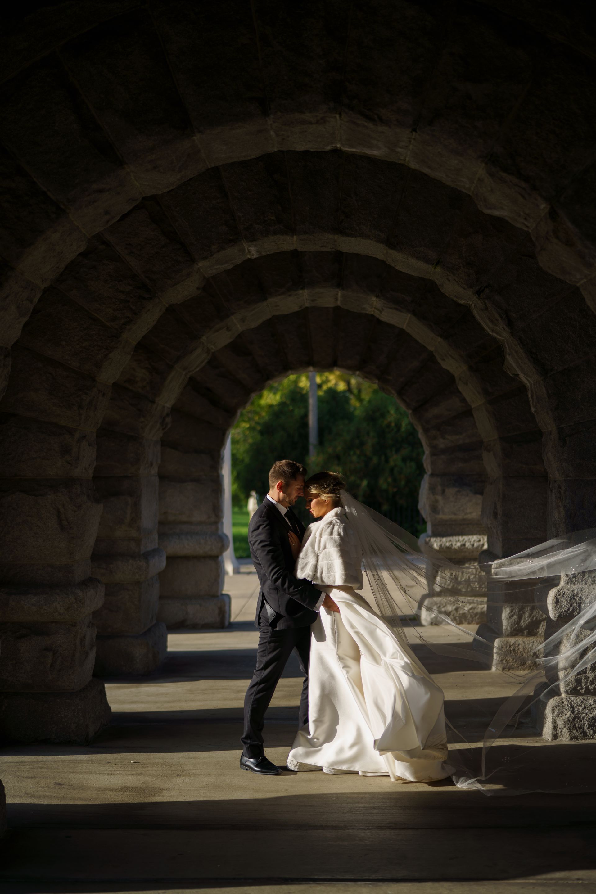 Couple embraces under stone archway; bride in white gown, groom in black suit. Sunlight filters through.