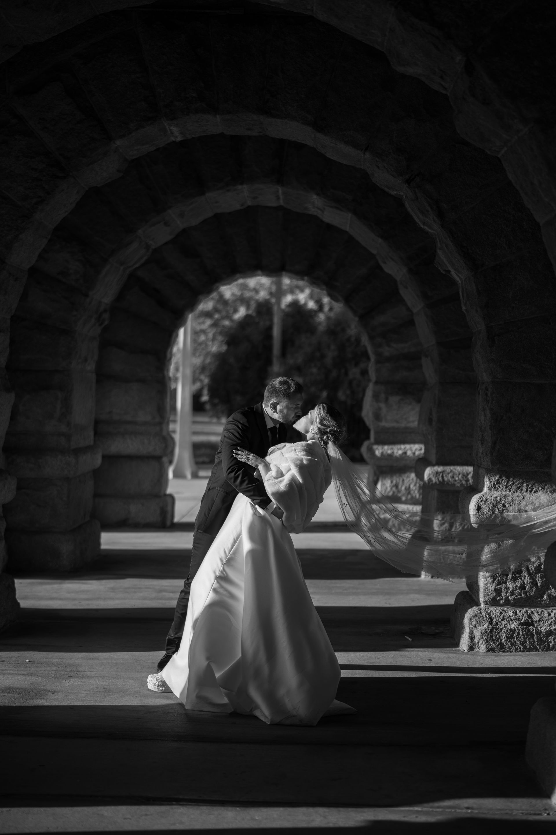 A couple in wedding attire dances under stone arches; black and white photograph.