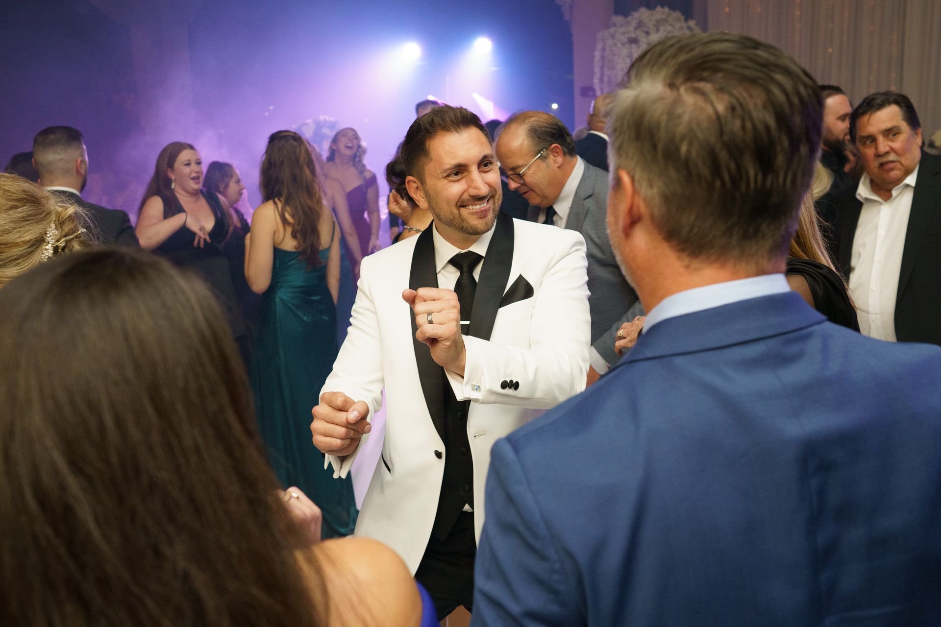 Man in white suit dances at a party; people in evening wear, colorful lights.