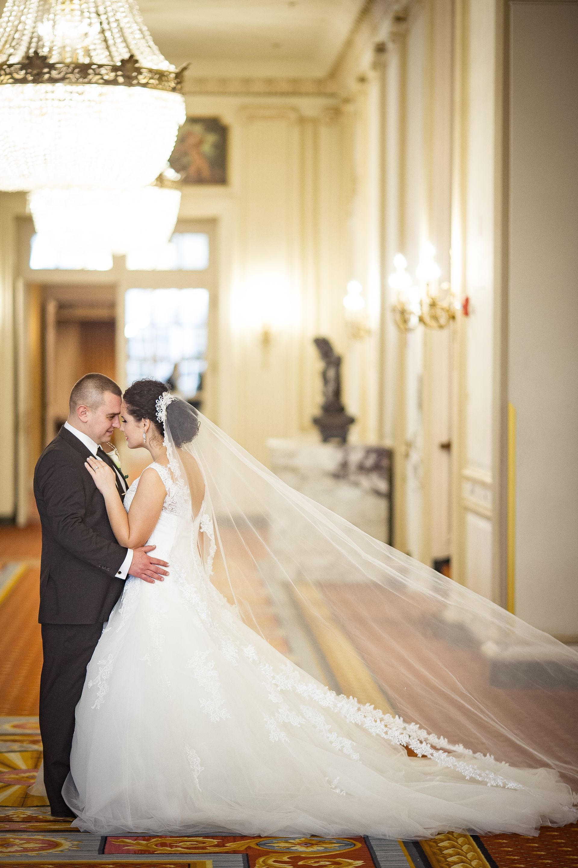 Bride and groom embrace in a formal hallway. The bride wears a white gown and veil, the groom, a dark suit.