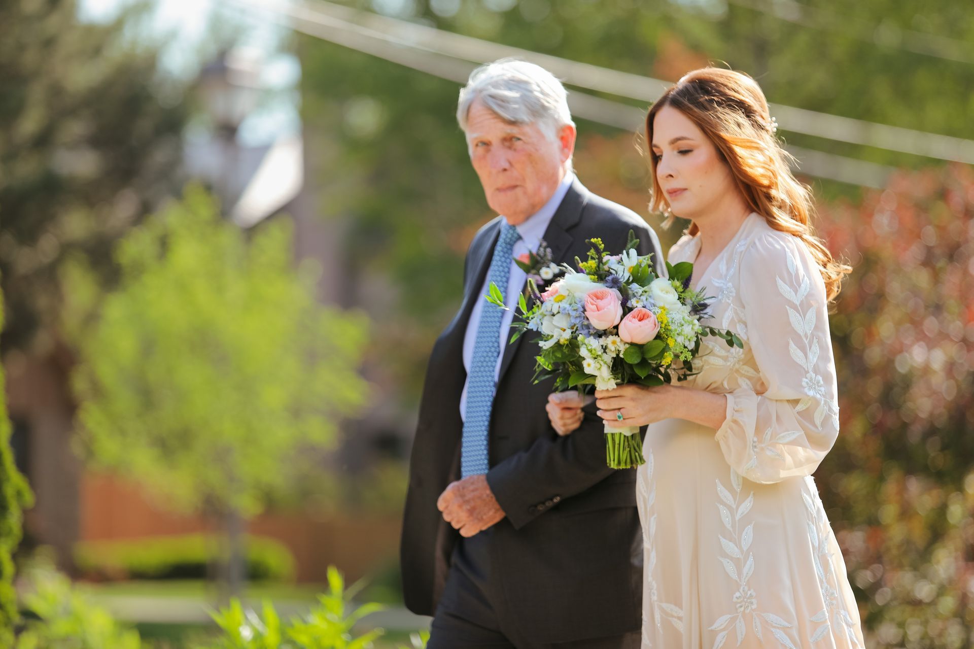 Bride walks with older man, arm in arm, towards ceremony. She wears a cream dress, he wears a suit.