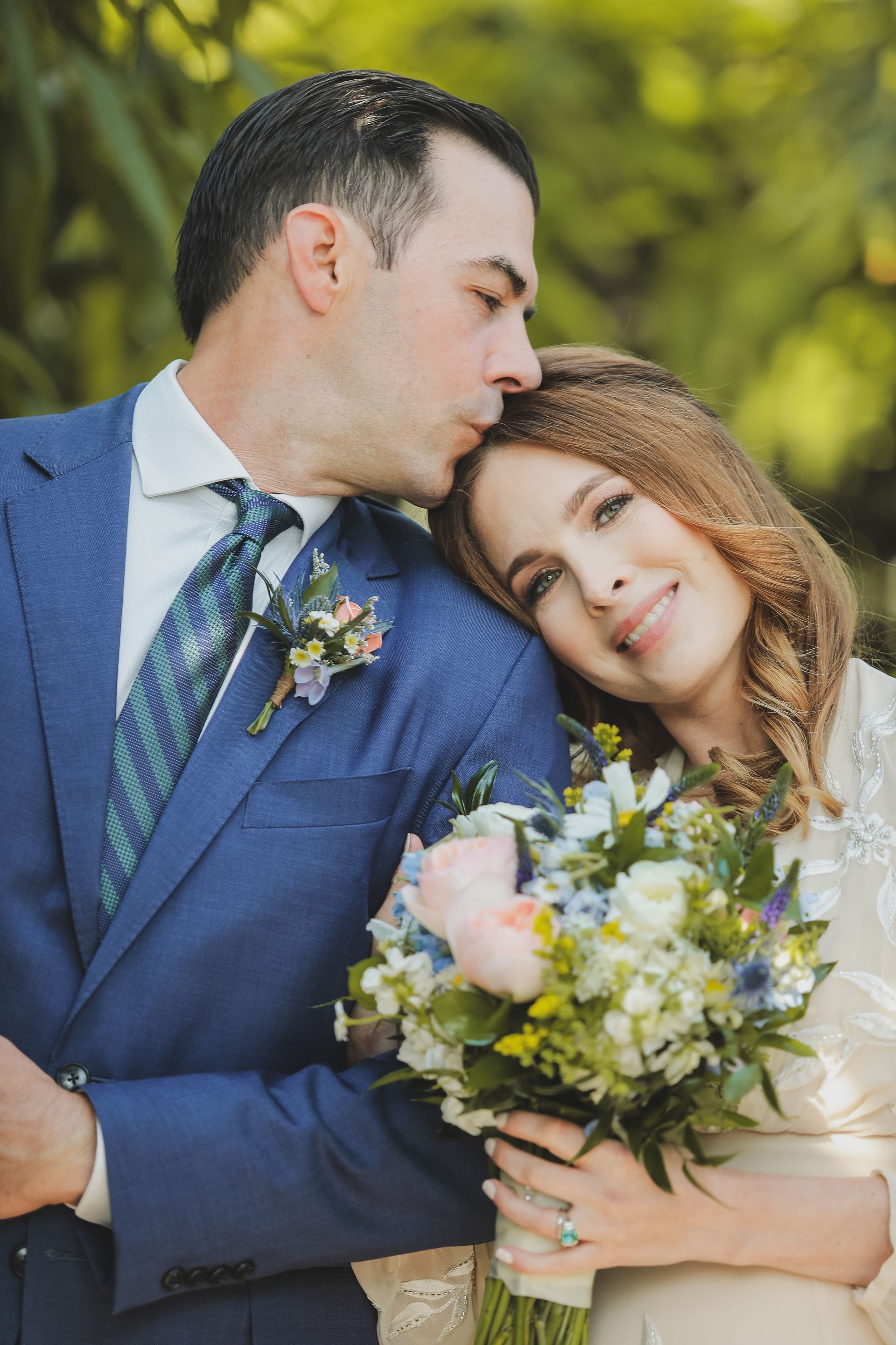 Groom in blue suit kisses bride's forehead. Bride holds bouquet, smiles. Outdoors.