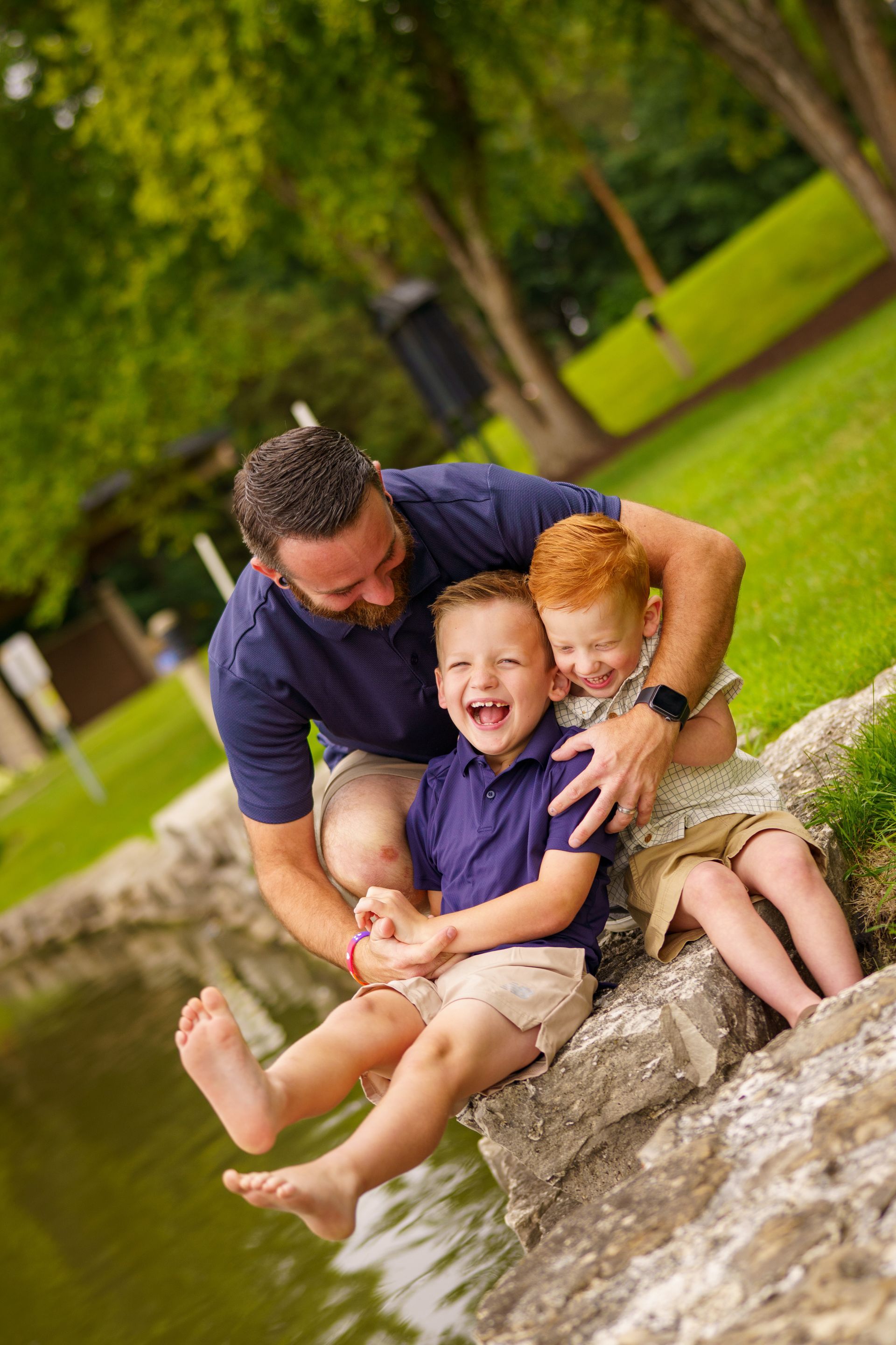 Man and two laughing children by water. Man hugs the boys, who sit on a rock. Green trees in background.