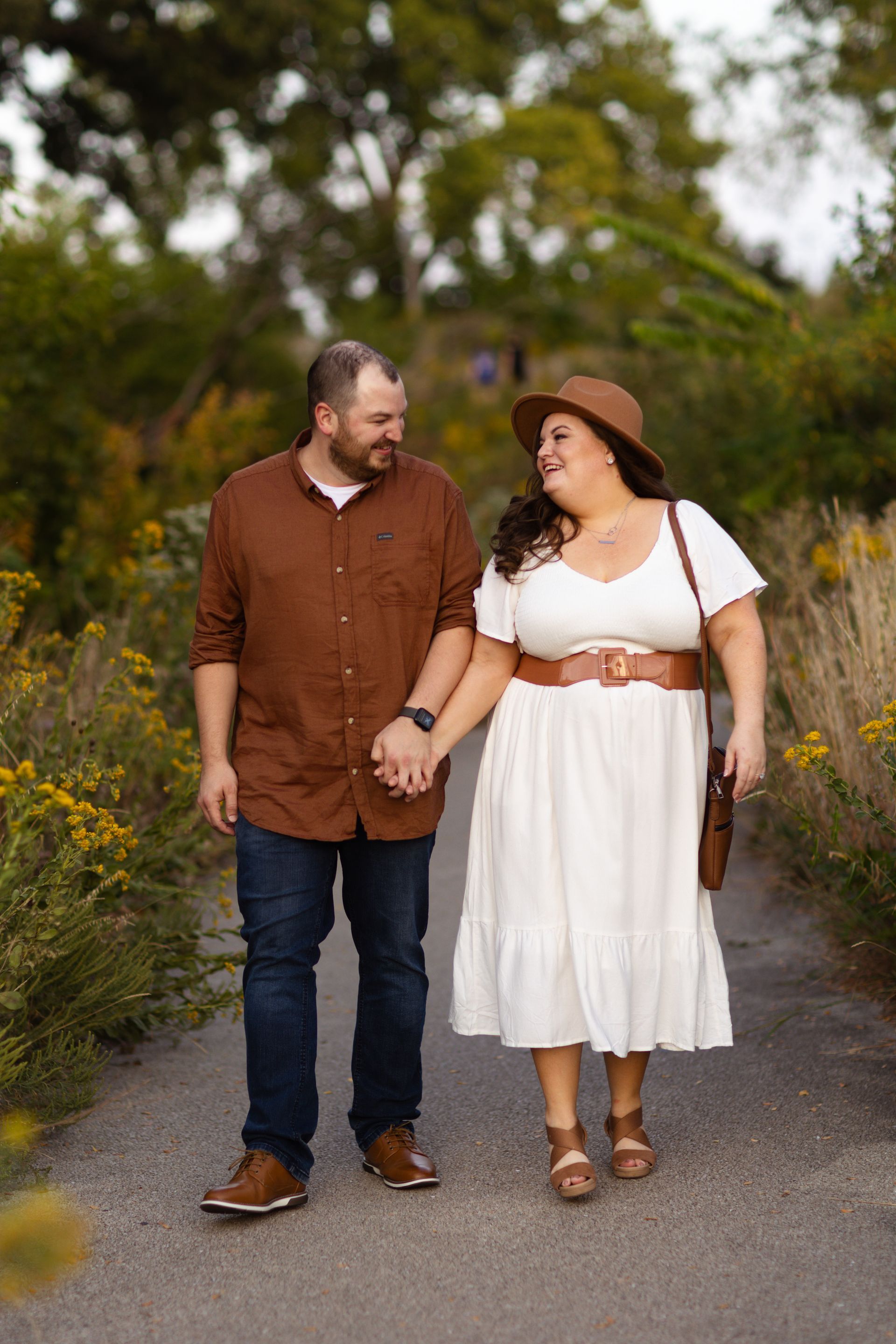 Couple holding hands, walking on a path, looking at each other. Man in brown shirt, jeans; woman in white dress, hat.