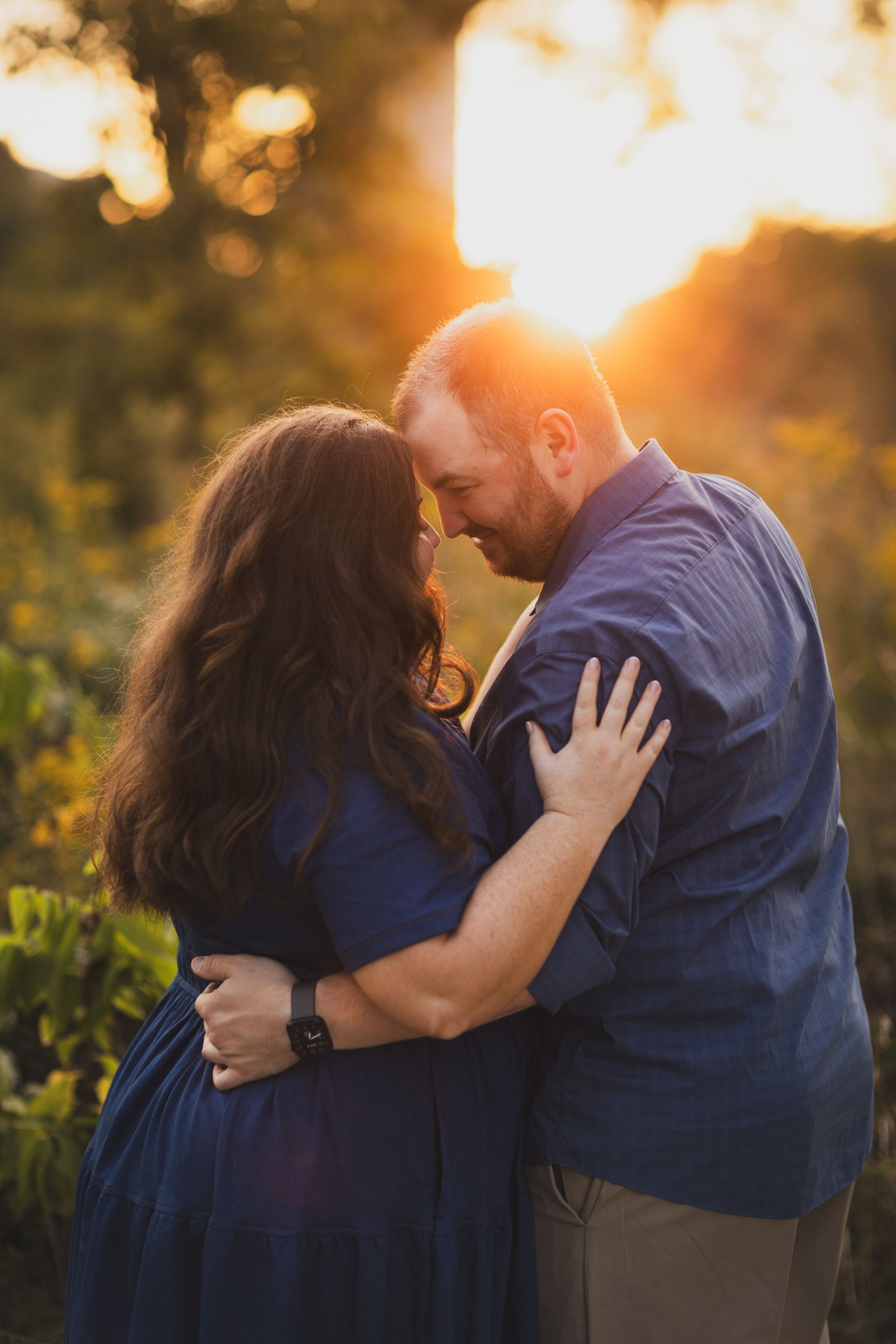 Couple embracing, heads touching, during sunset in a field; golden light, blue clothing.