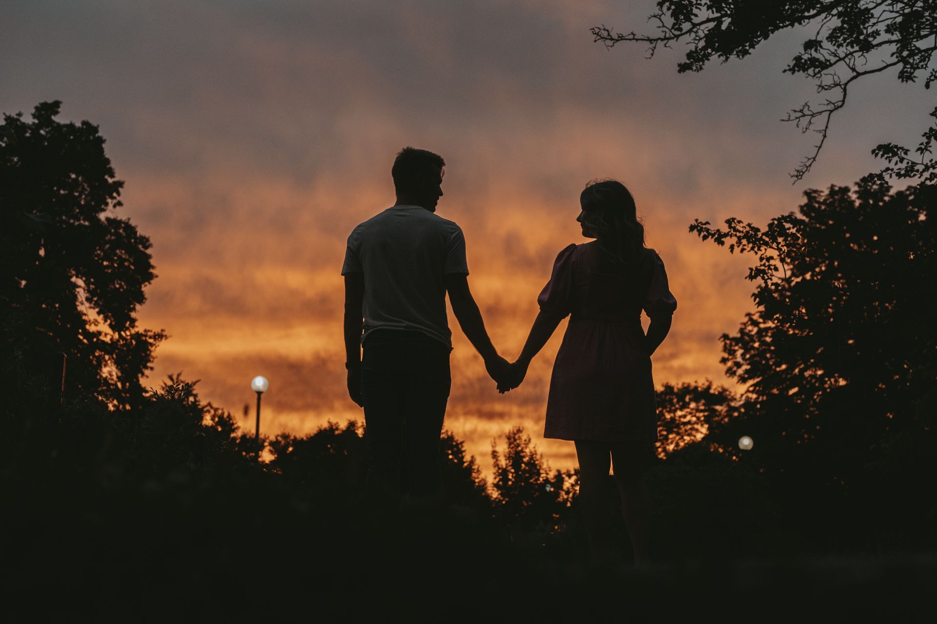Couple holding hands, silhouetted against a vibrant orange sunset. Trees frame the scene.