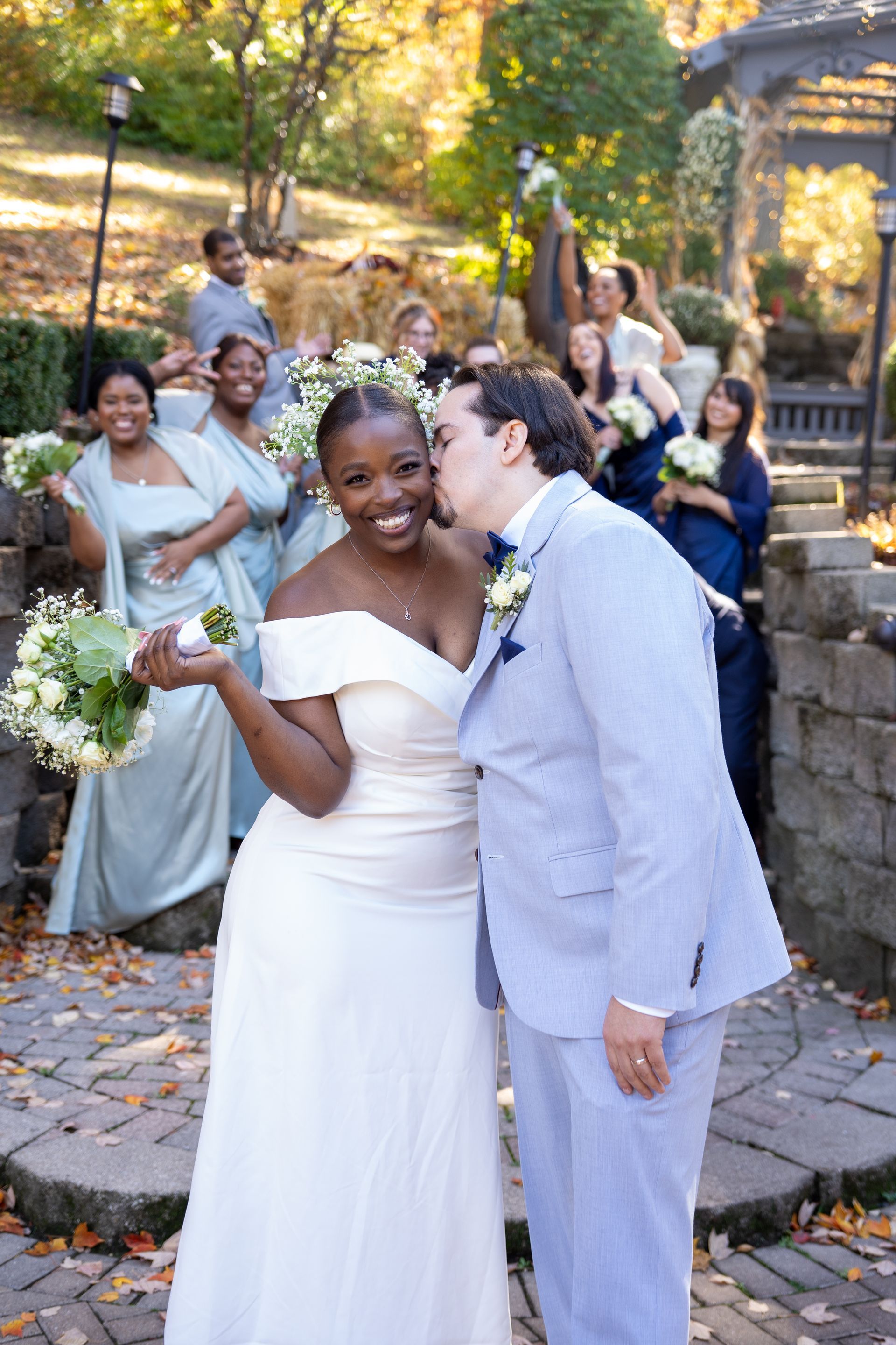 Bride and groom smile, groom kisses bride's cheek. Wedding party in background, stone steps, outdoor setting.