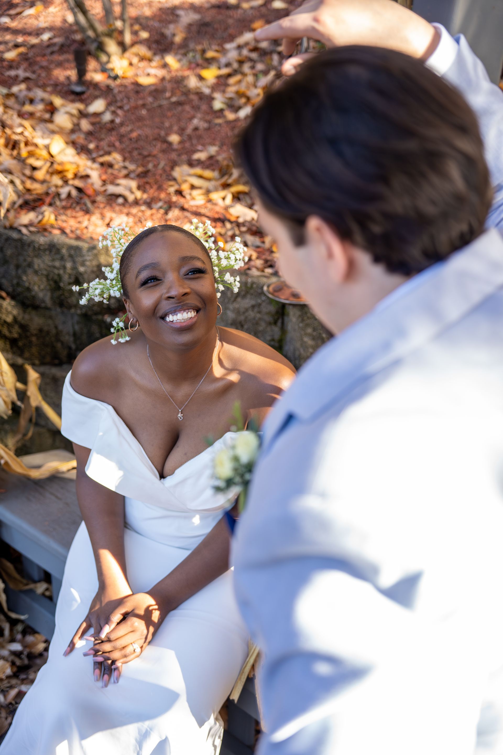 Bride in off-the-shoulder white dress smiles up at person, possibly groom, outdoors.