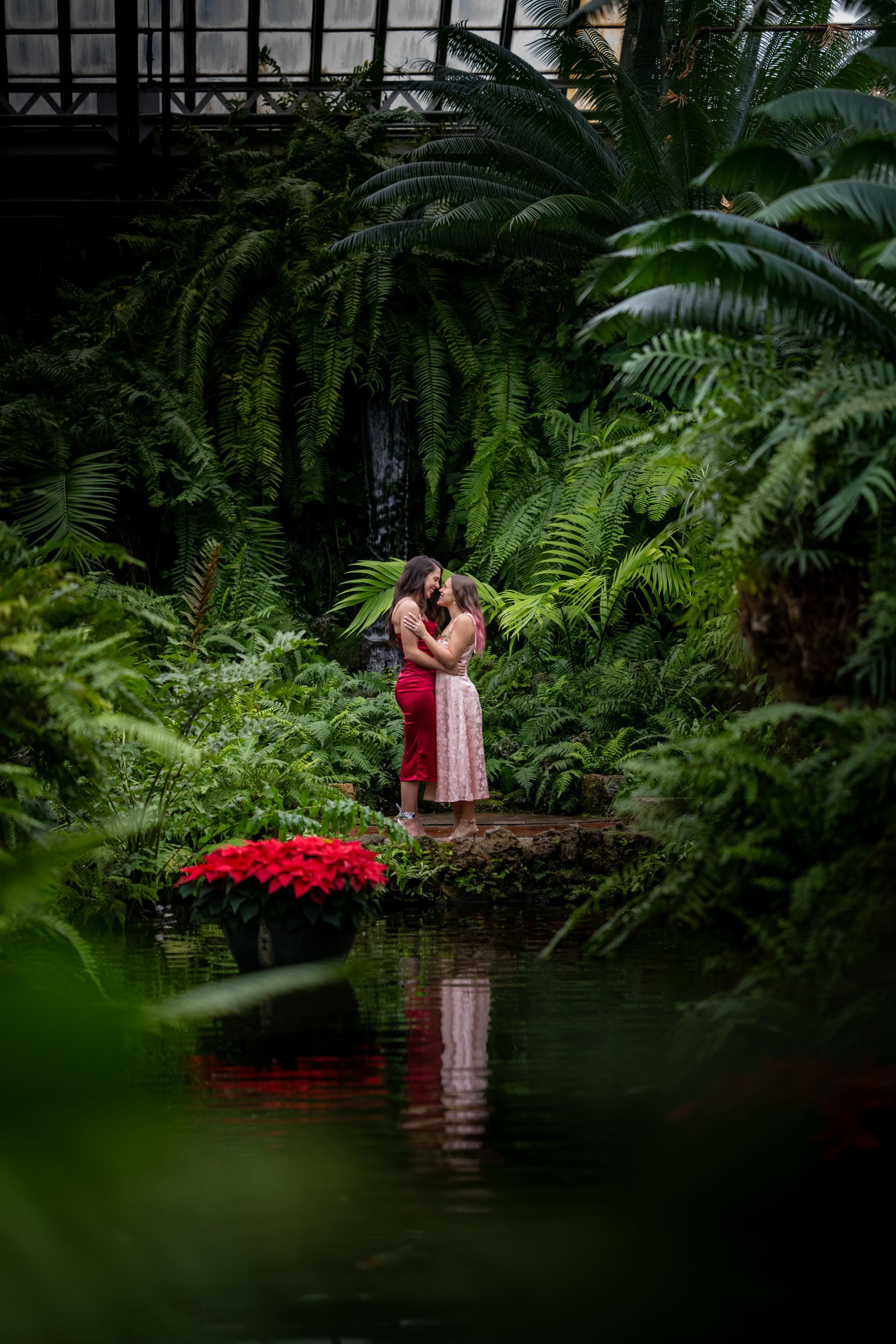 Two people embrace by a pond in a lush, green greenhouse. One wears a red dress, the other a pink one.