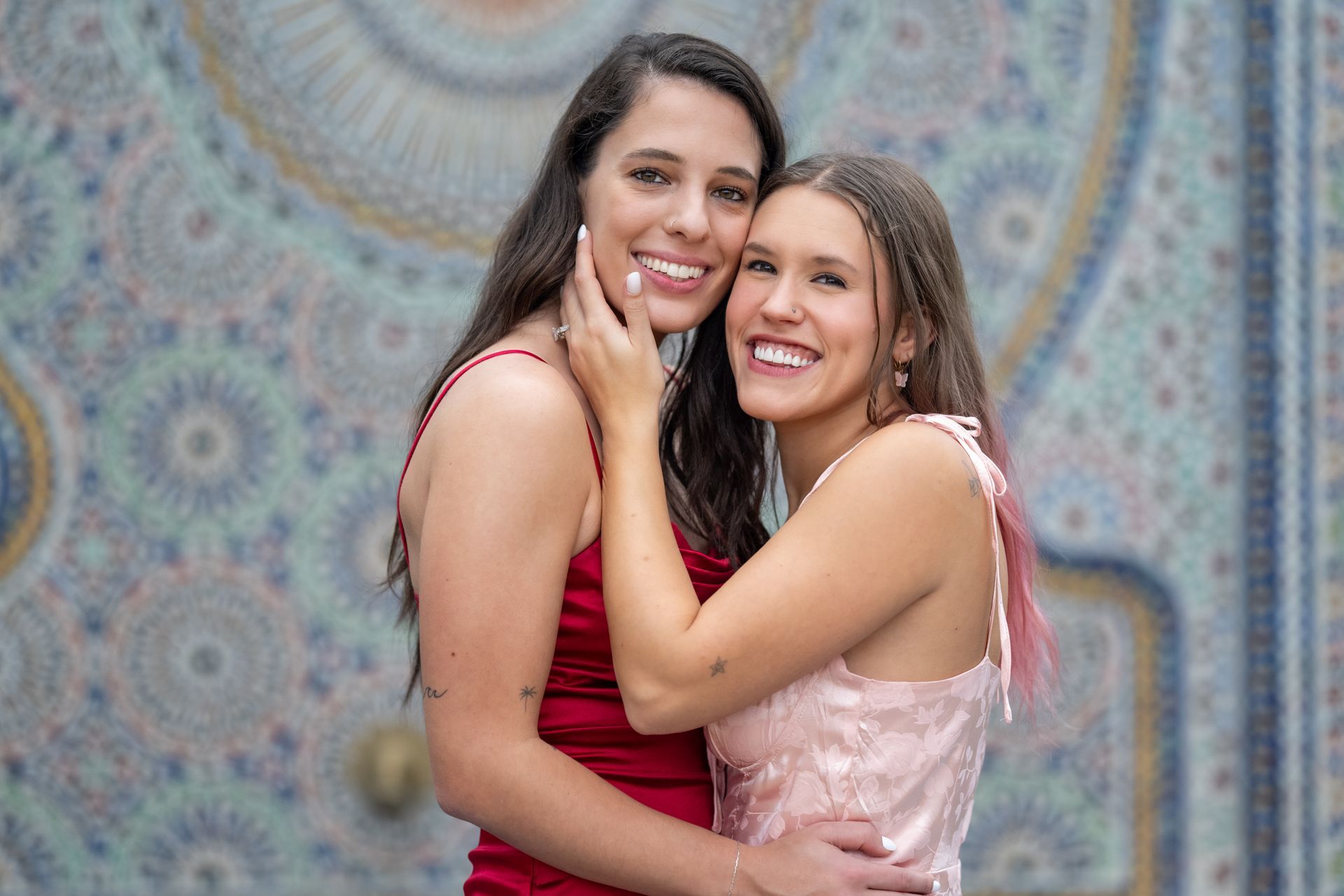 Two smiling women embrace; one in red, the other in pink, in front of a patterned wall.
