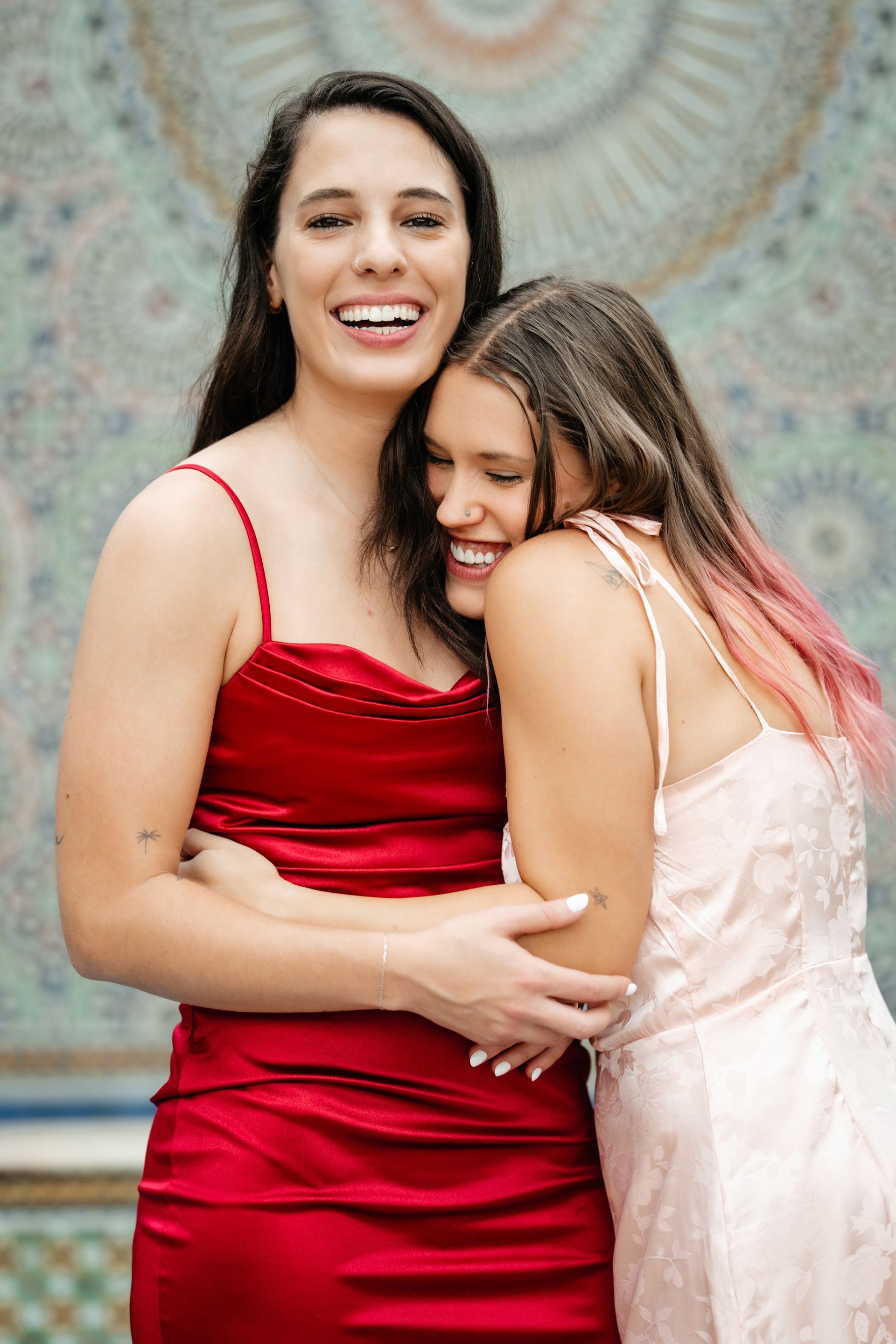 Two women hugging and smiling; one in a red dress, the other in a light pink dress, against a tiled wall.
