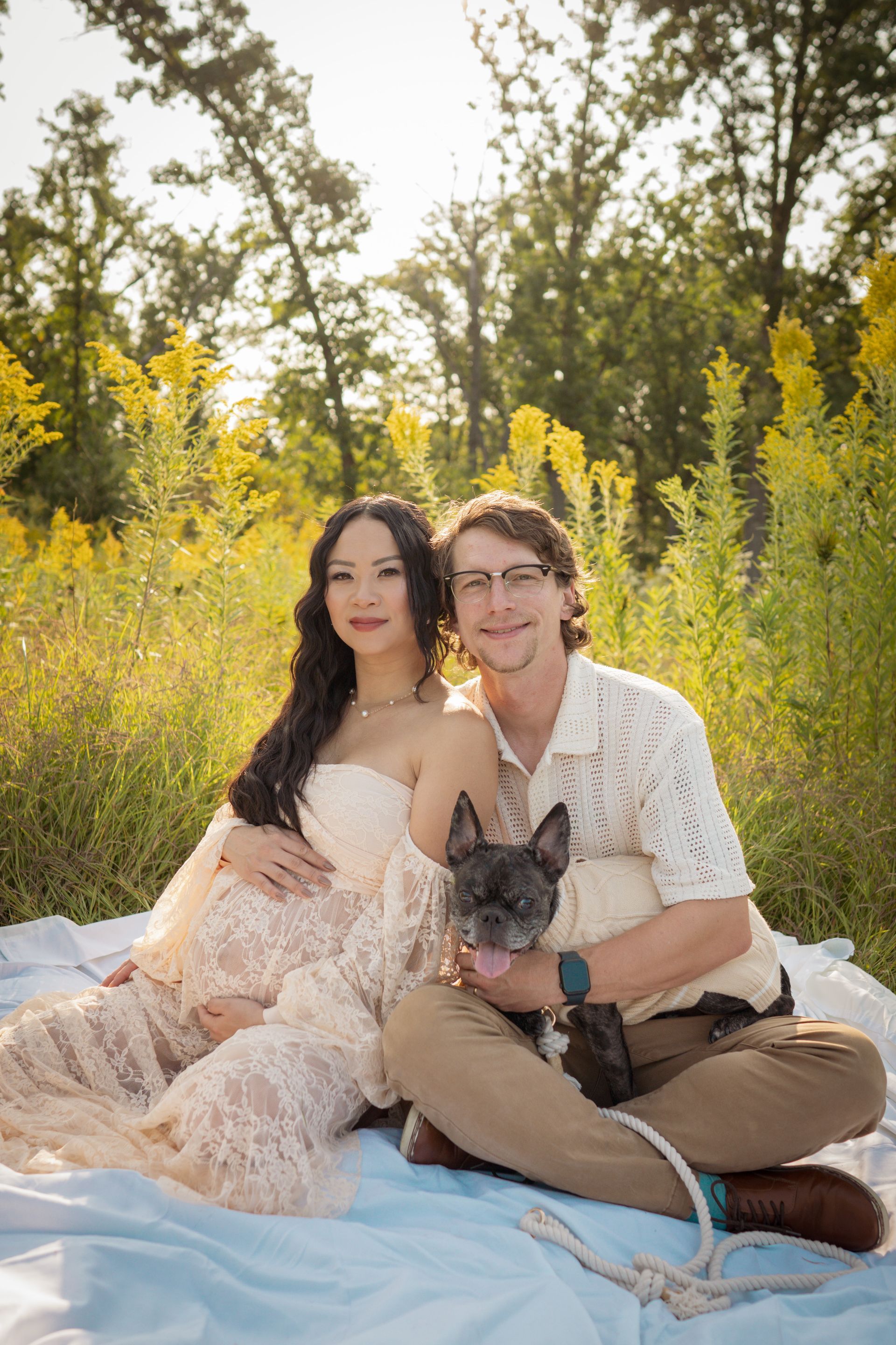 Couple with French bulldog on a blanket outdoors in front of yellow flowers; woman is pregnant.