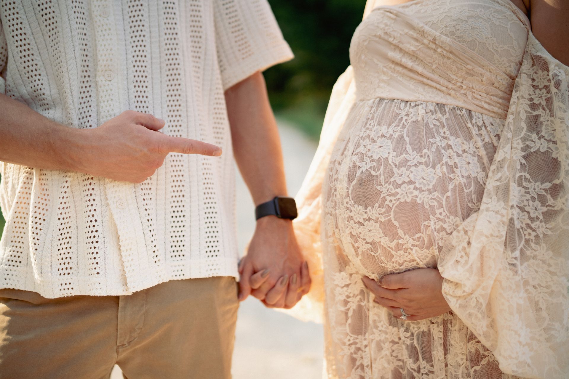 A pregnant person in a cream lace dress holds hands with a person in a white shirt and khaki pants, who points.