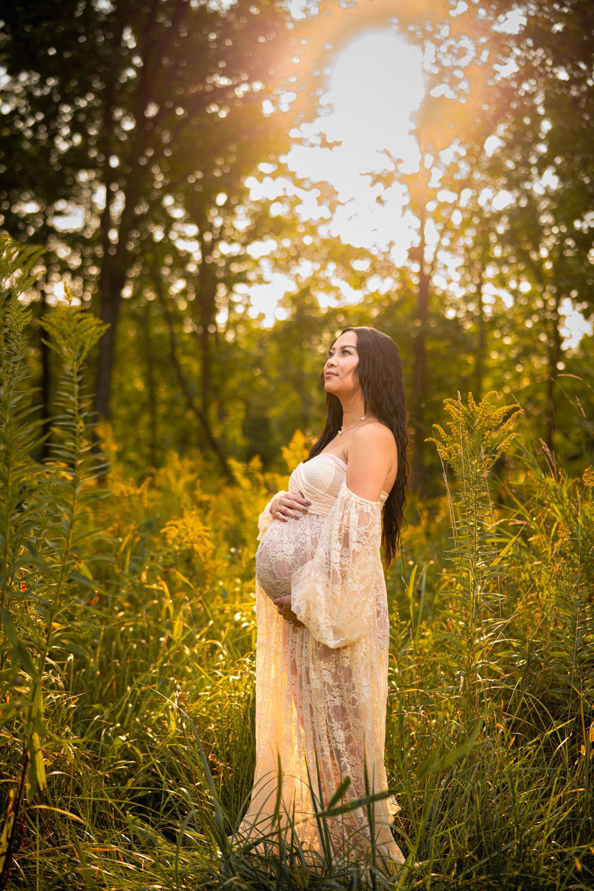 Pregnant person in a flowing gown stands in a field of tall grass, bathed in sunlight.