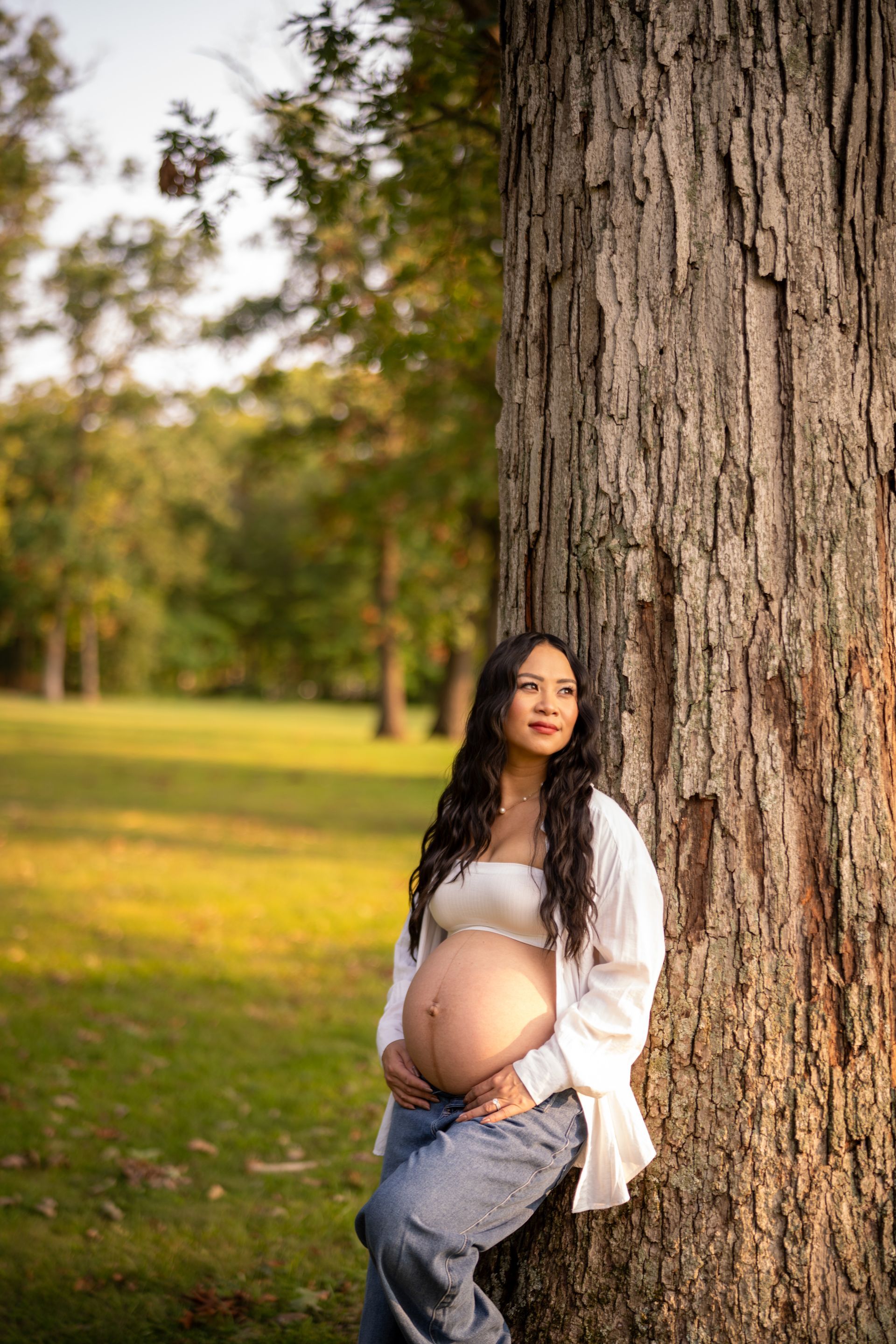 Pregnant woman leans against a tree, hand on belly, wearing white top and jeans. Outdoors, sunny.