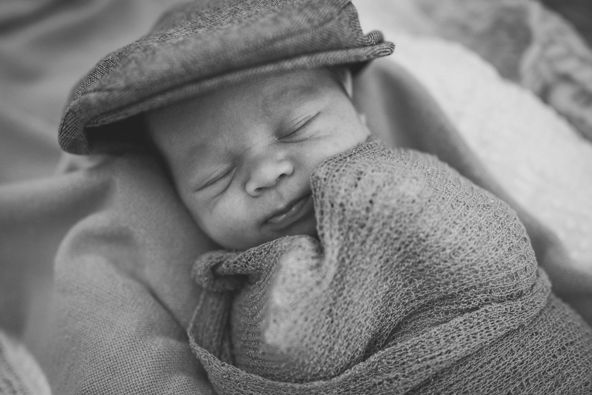 Newborn baby sleeps, wrapped in a blanket, wearing a newsboy cap.