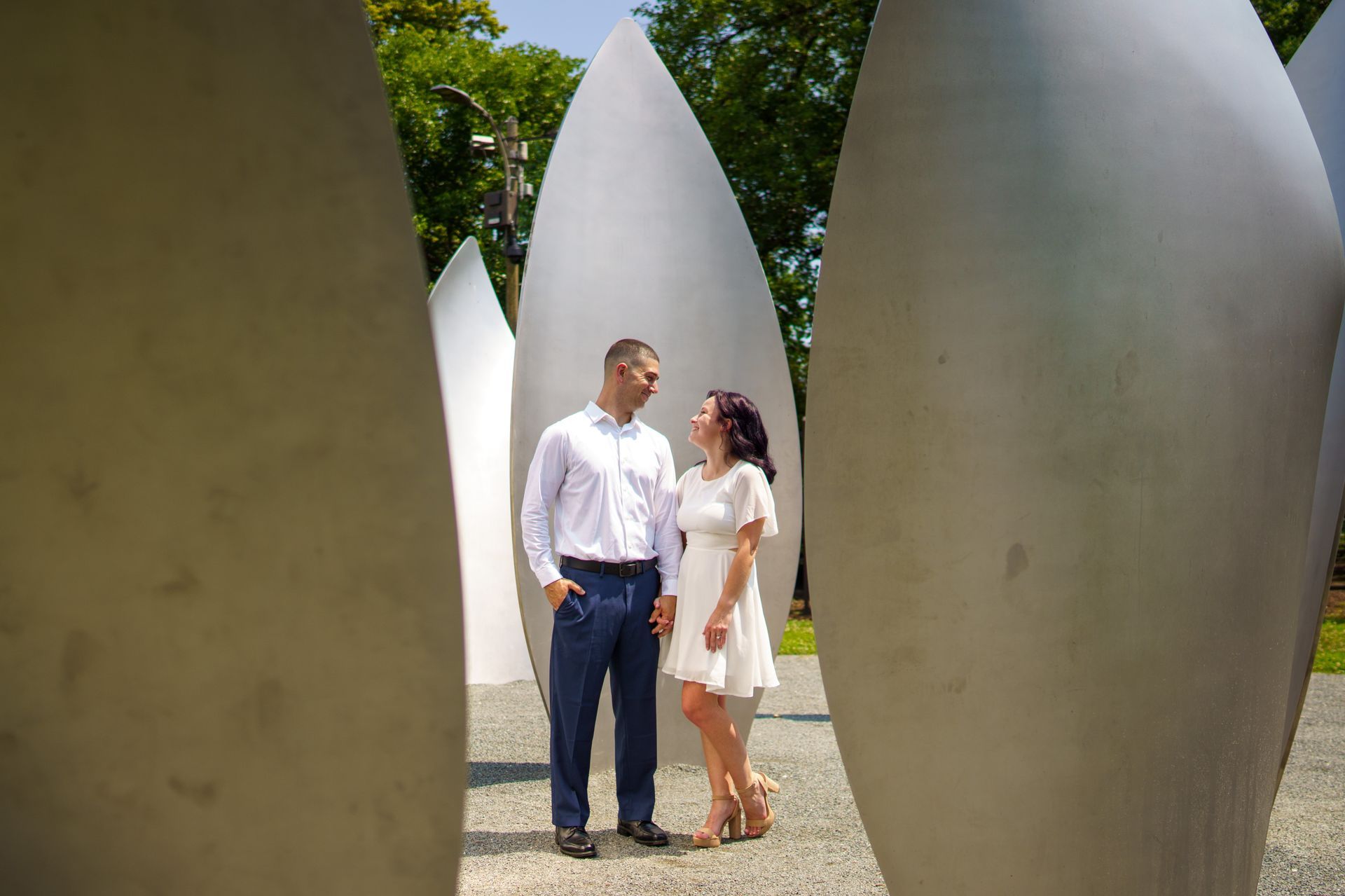Couple standing, looking at each other, between large silver sculptures in an outdoor setting on a sunny day.