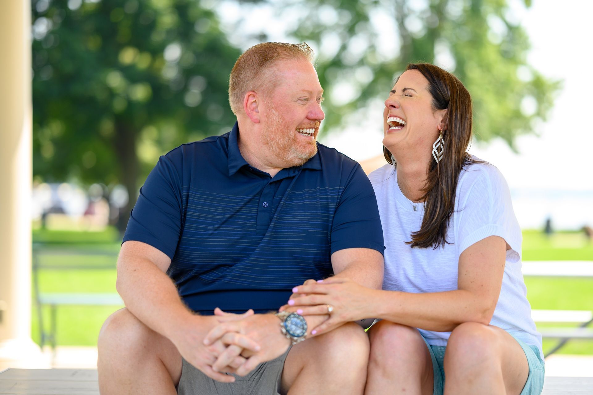 Couple laughing together, sitting under a gazebo. Man in blue shirt, woman in white shirt. Outdoors in the sun.