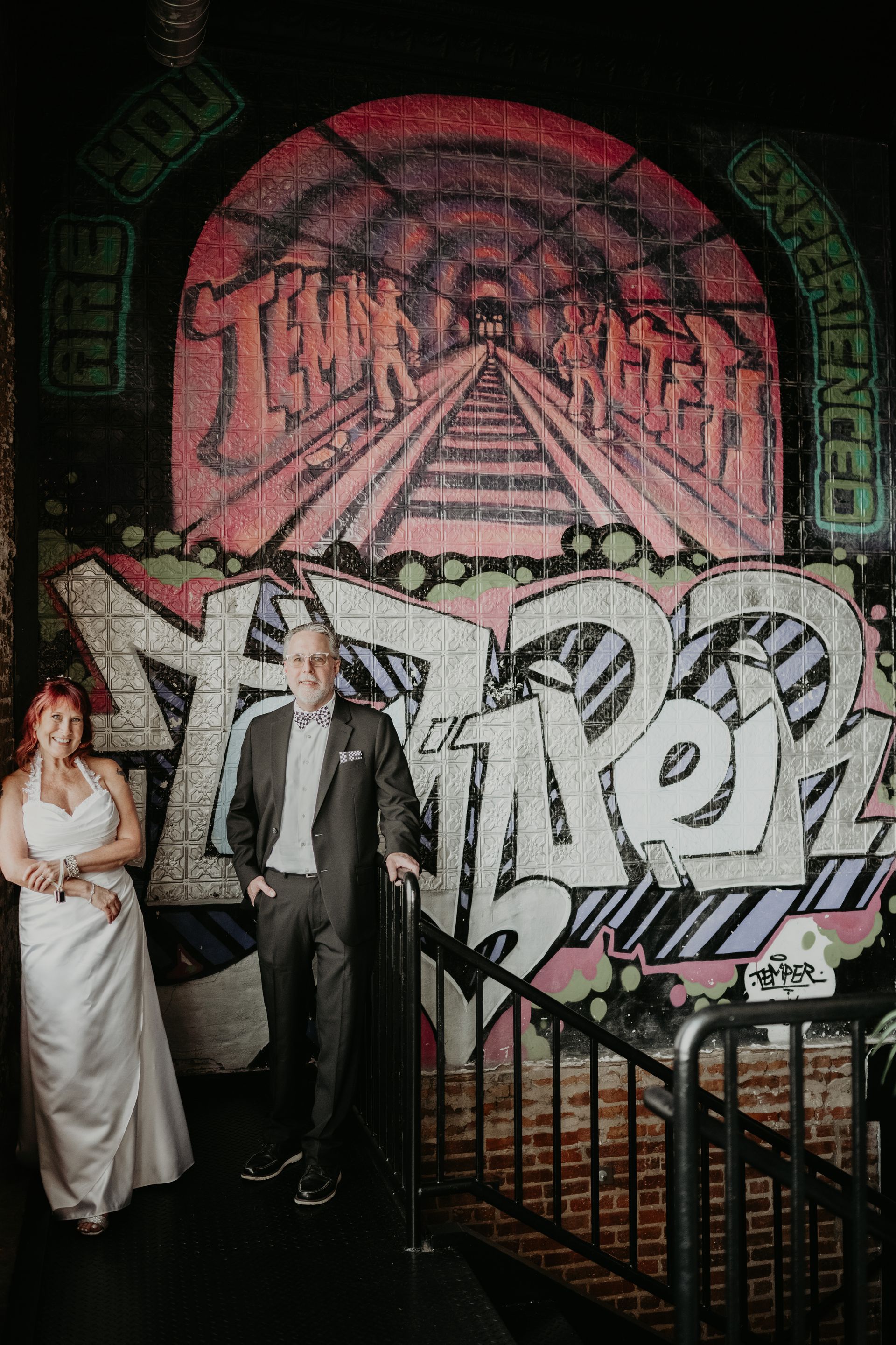 Couple in wedding attire posing by graffiti art with a train tunnel image.