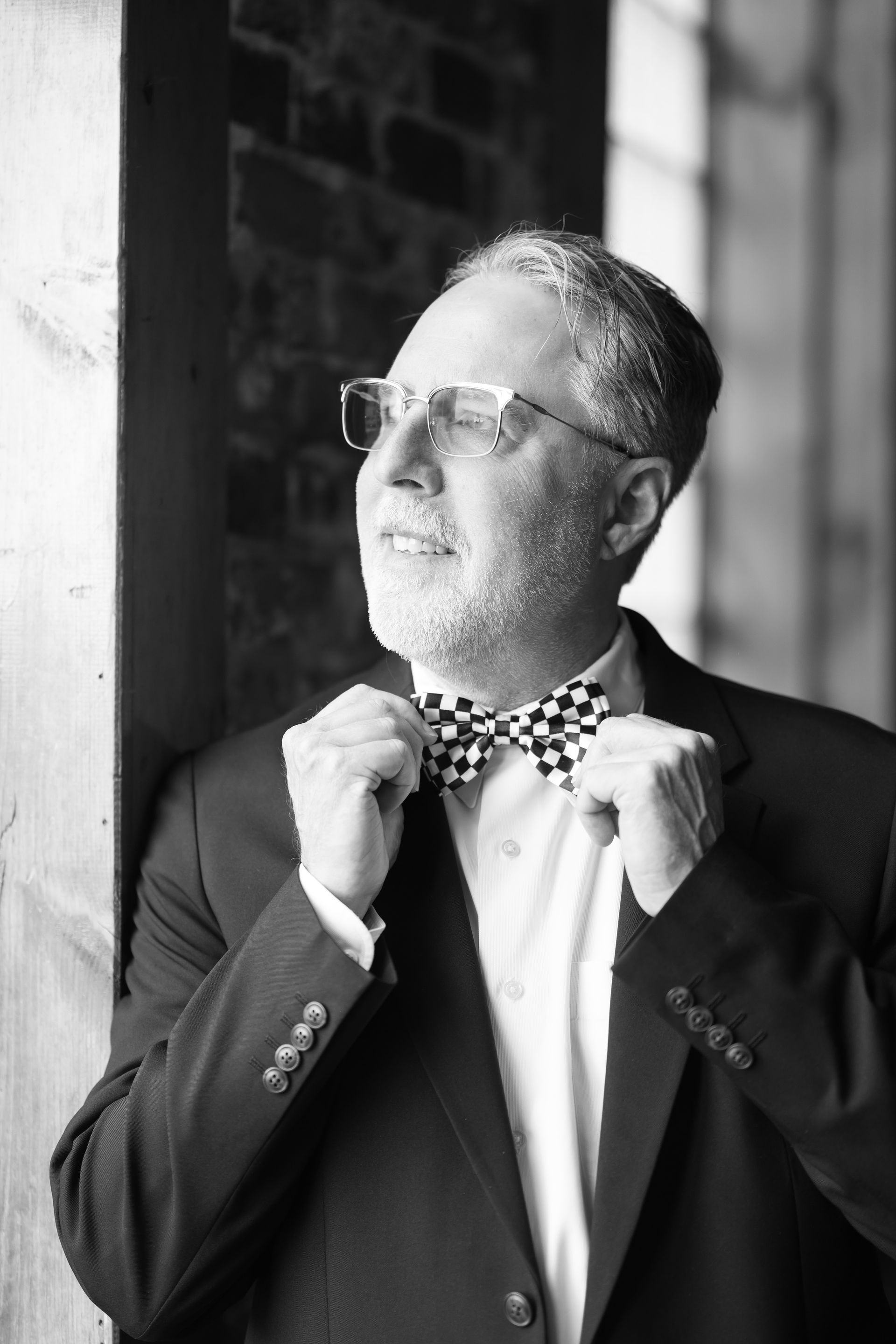 Man in suit adjusts checkered bow tie, looking towards the window in a brick building.