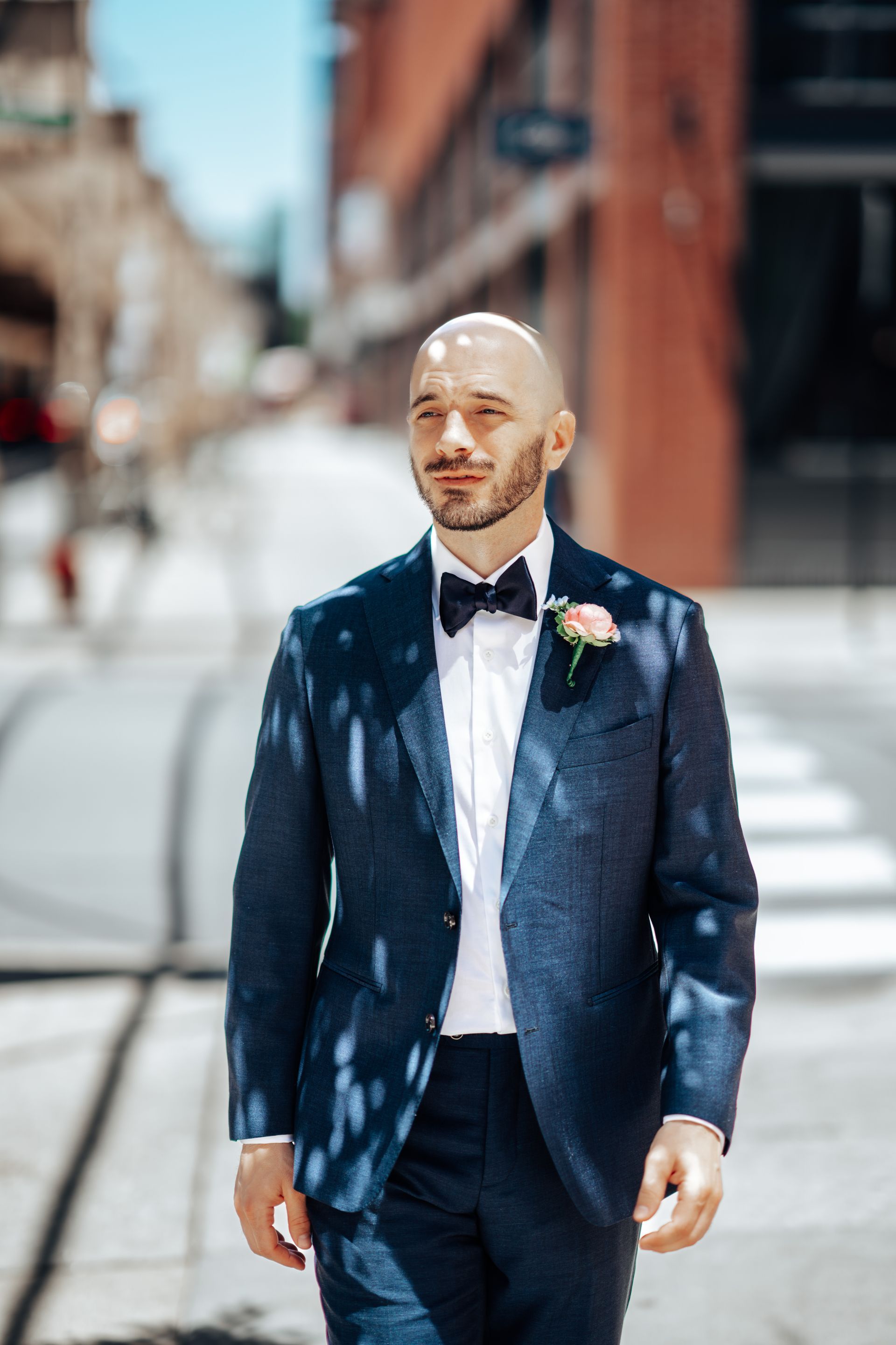 Man in a navy blue suit walks down a city street, wearing a bow tie and a flower.
