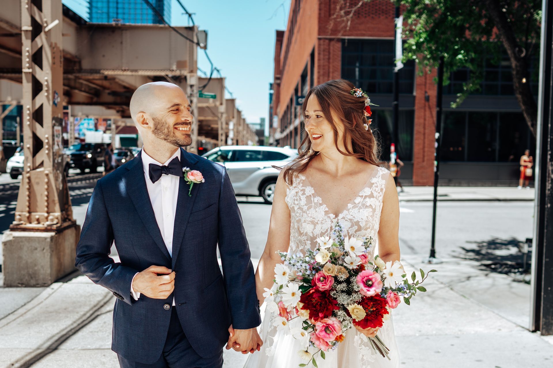 Bride and groom walking, holding hands, smiling, in city; she carries flowers.