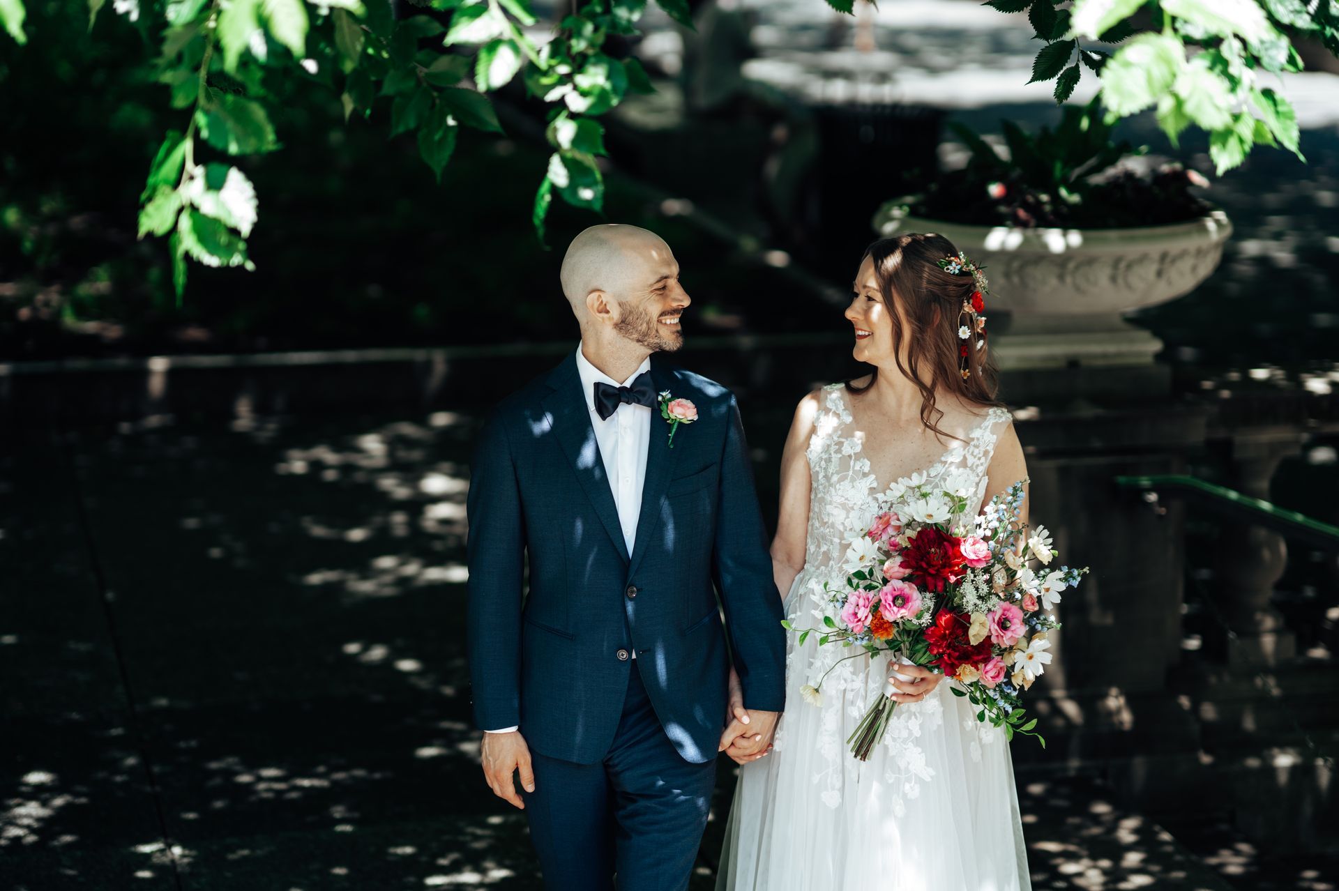 Couple holding hands, smiling at each other, in a sunlit garden, bride holding bouquet, groom in a blue suit.