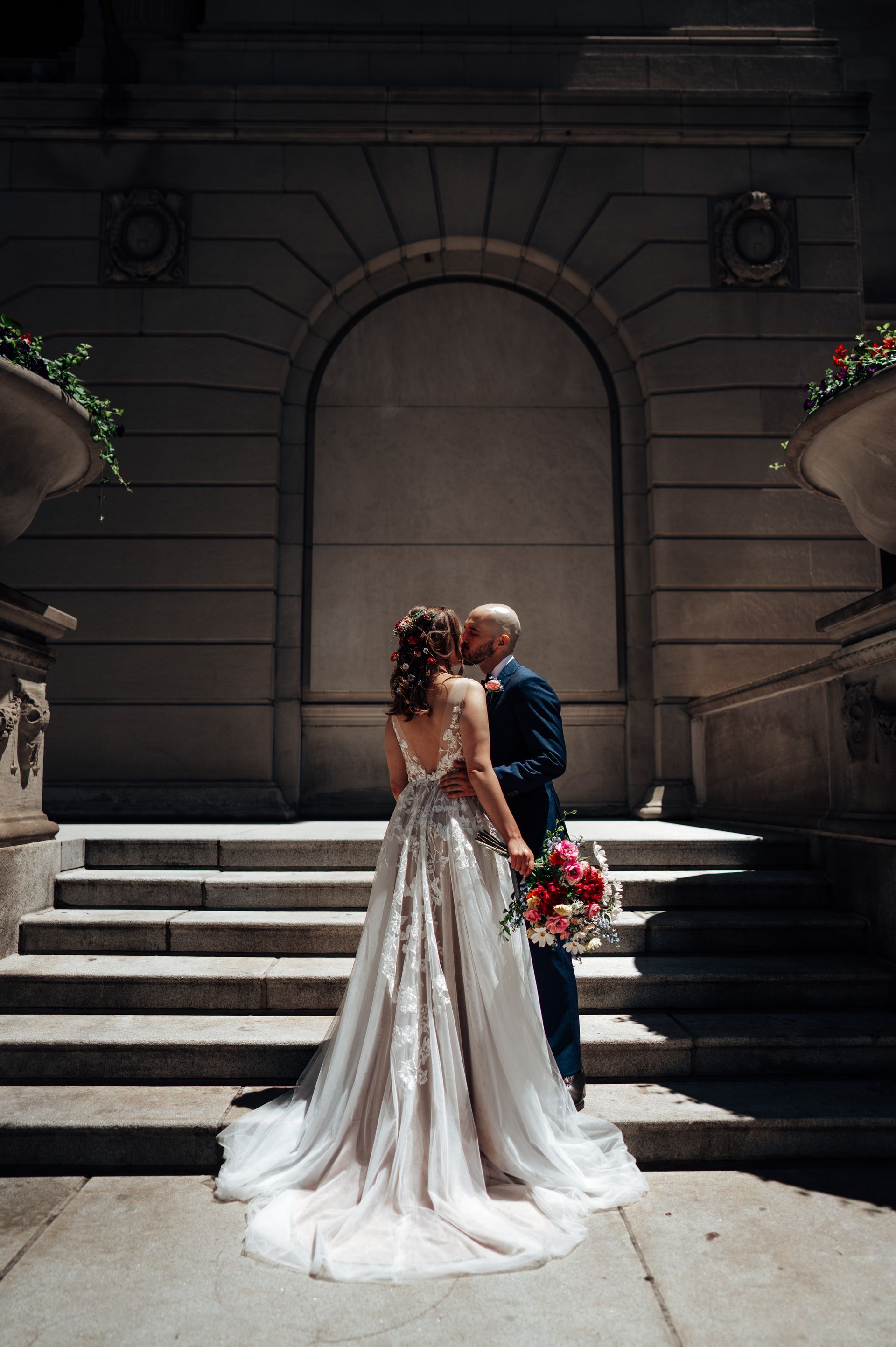Couple kissing on steps in front of a stone building with a large arched entrance; the bride wears a flowing gown.