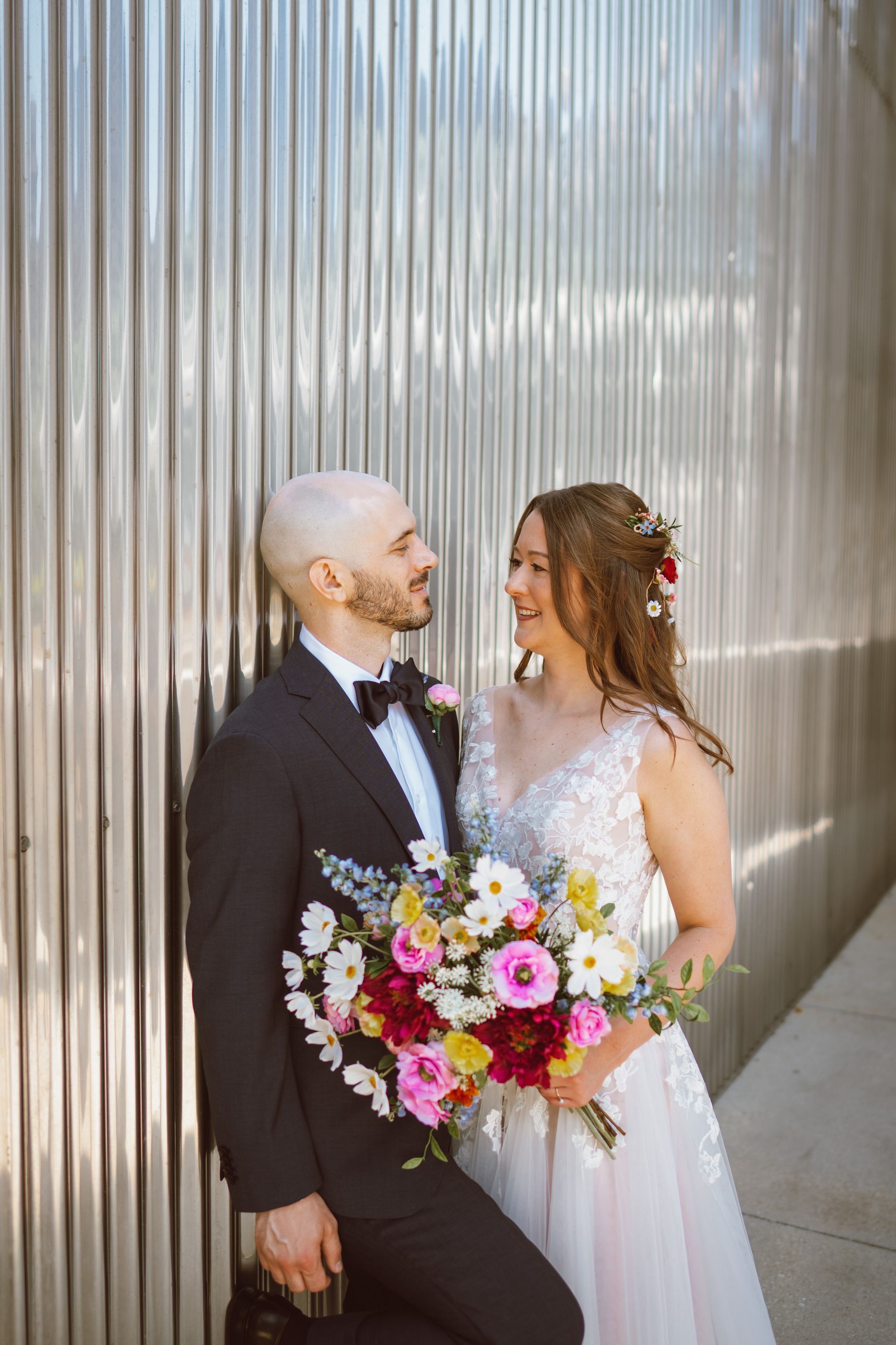 Wedding couple smiles, holding a colorful bouquet, against a ridged silver wall.
