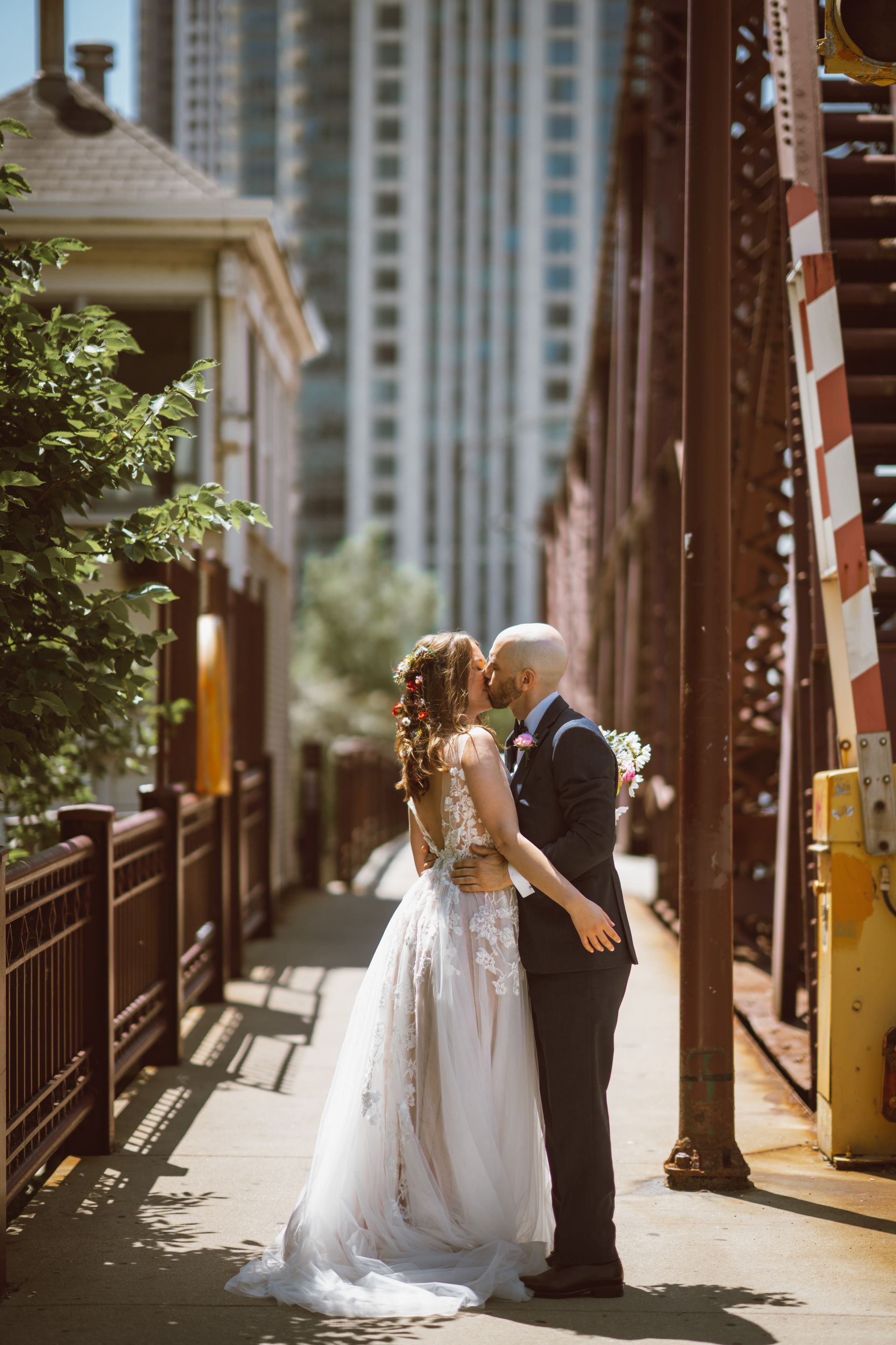 Wedding couple kissing on a metal bridge, city buildings in the background. Bride in white gown; groom in dark suit.