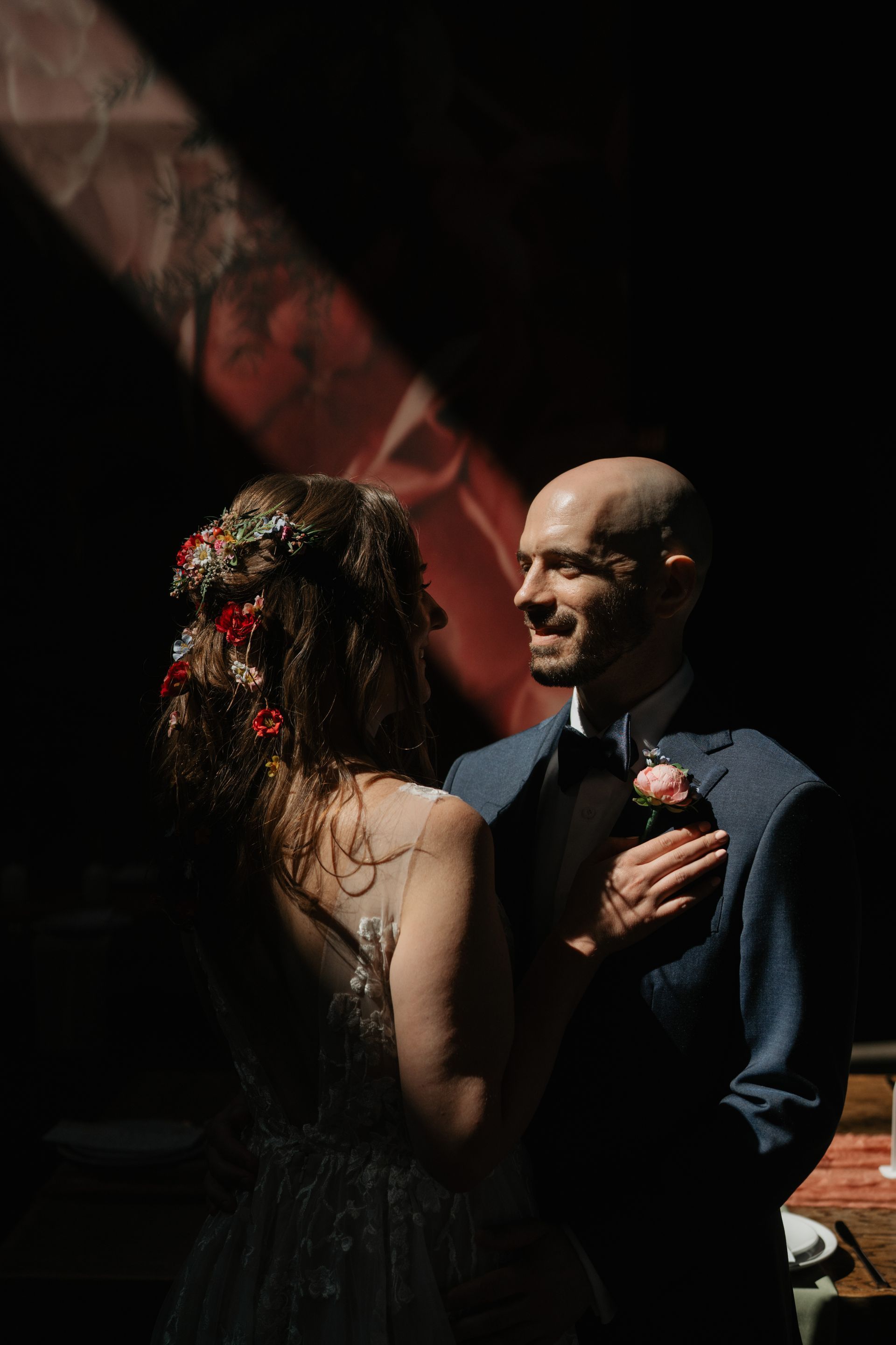 Couple gazing at each other. Bride with floral hairpiece, groom in blue suit. Soft light on faces, dark background.