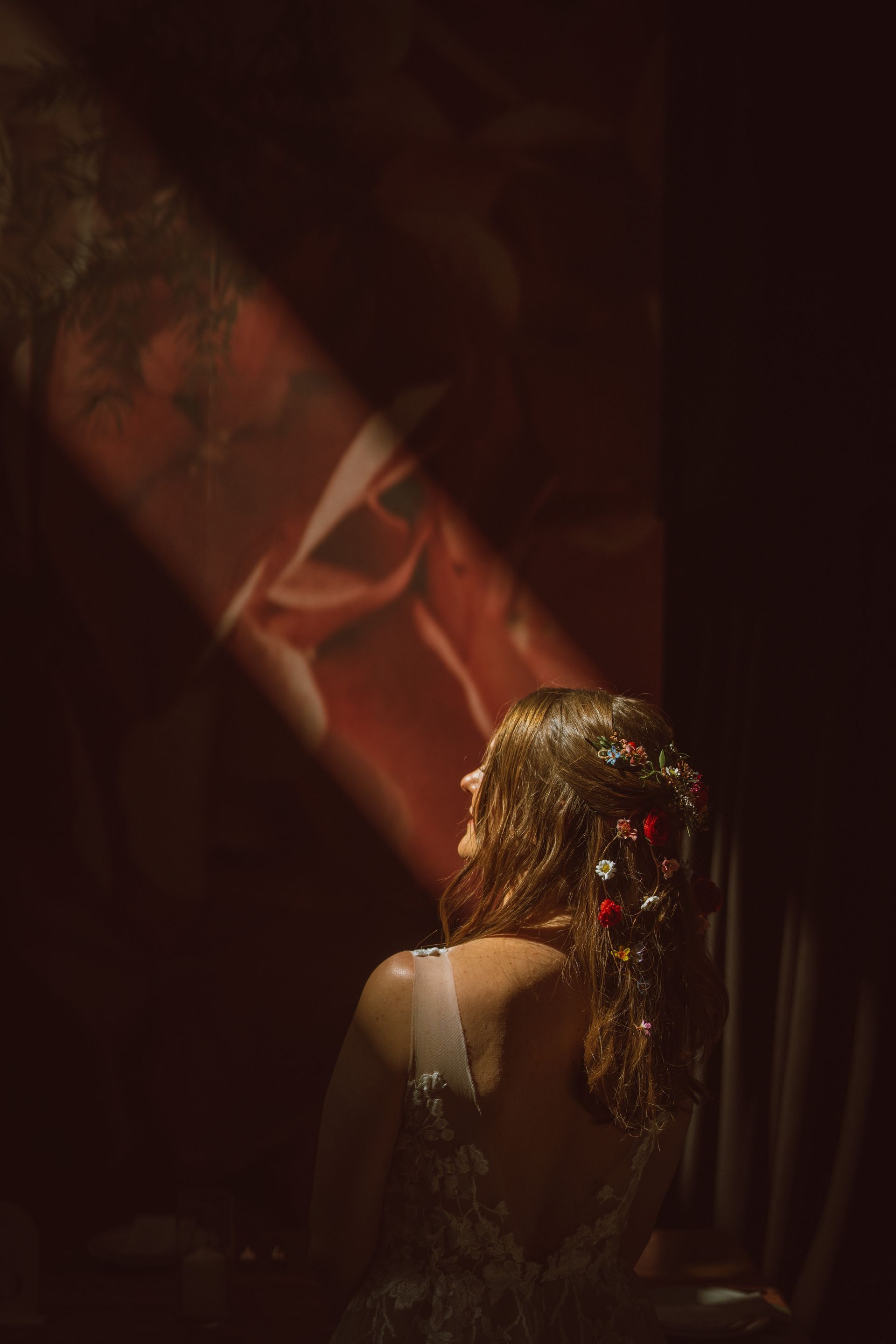 Woman with flower-adorned hair, lit by a beam of light, standing before a red wall.