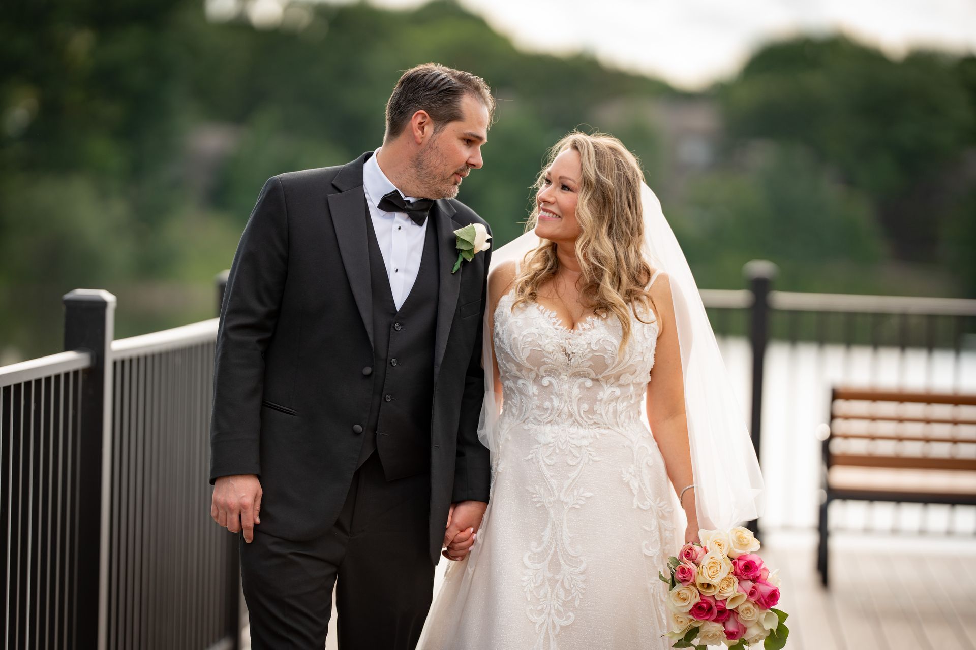Bride and groom holding hands, smiling at each other on a wooden dock; water in background.