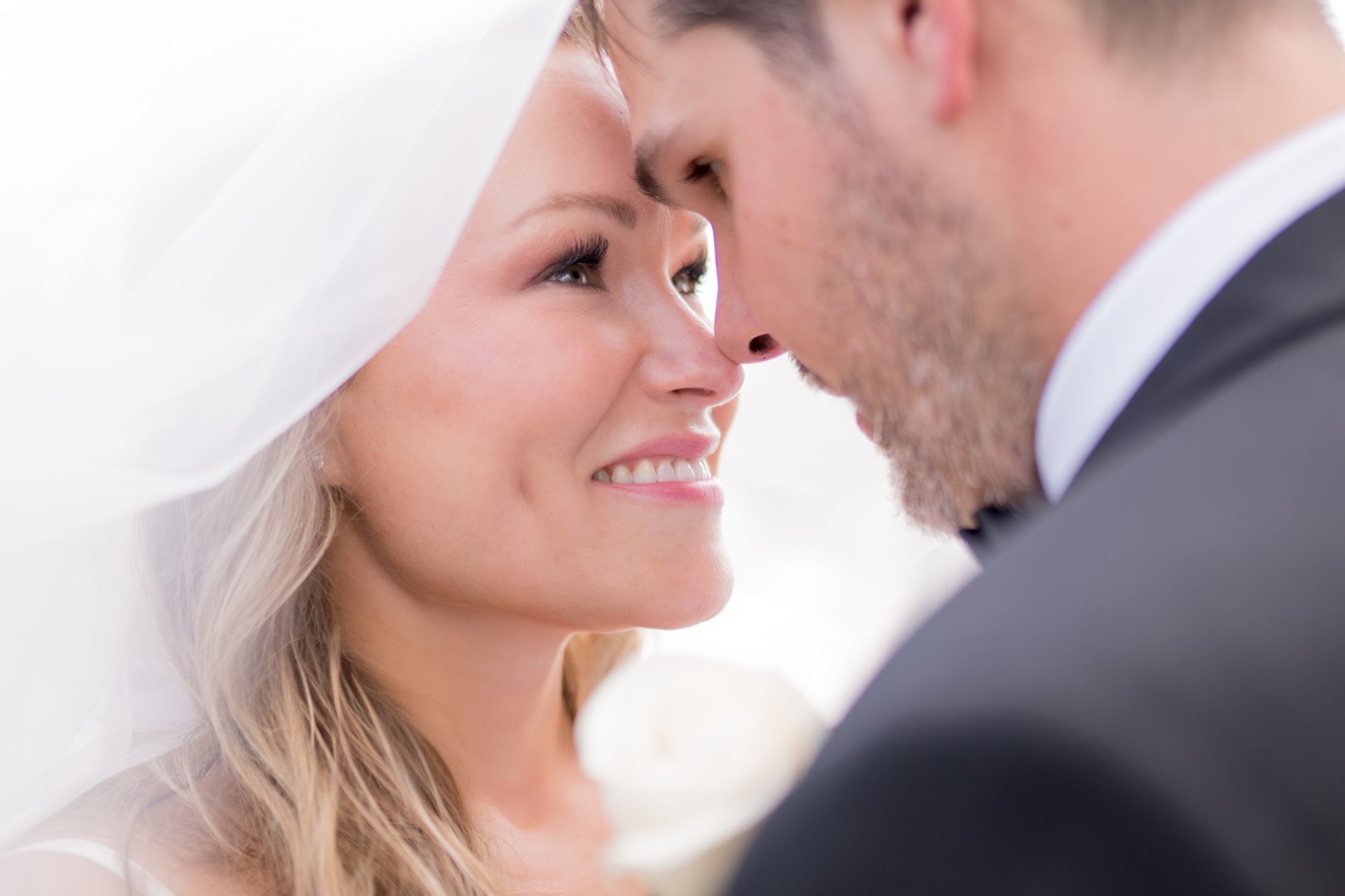 Bride and groom face each other, touching noses, smiling. Bride wears veil, groom in suit.
