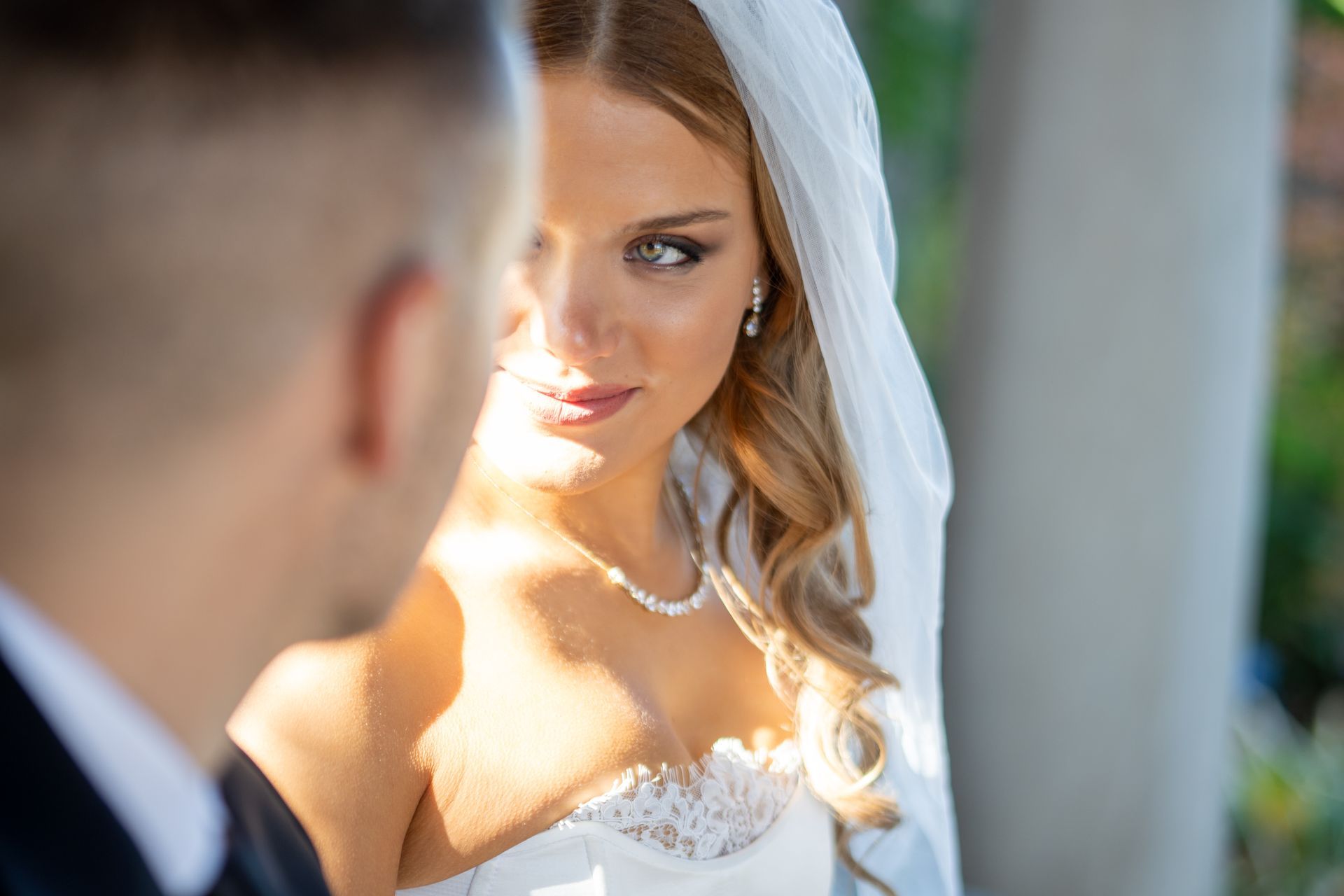 Bride in a white dress, veil, and jewelry looks at groom. Sunny outdoor setting.