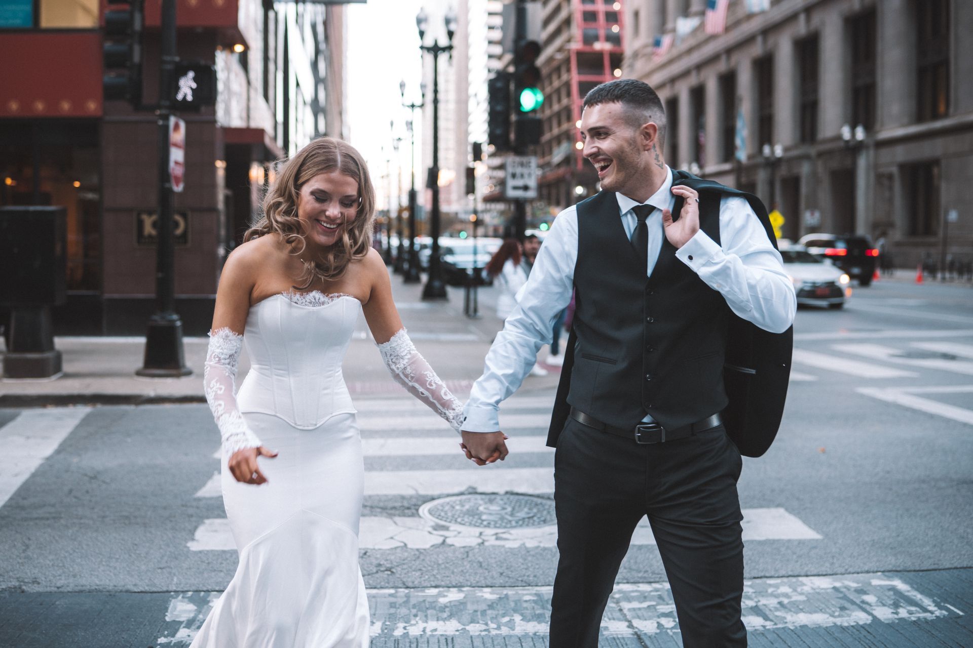Bride and groom holding hands, smiling, crossing a city street; the groom carries his jacket.