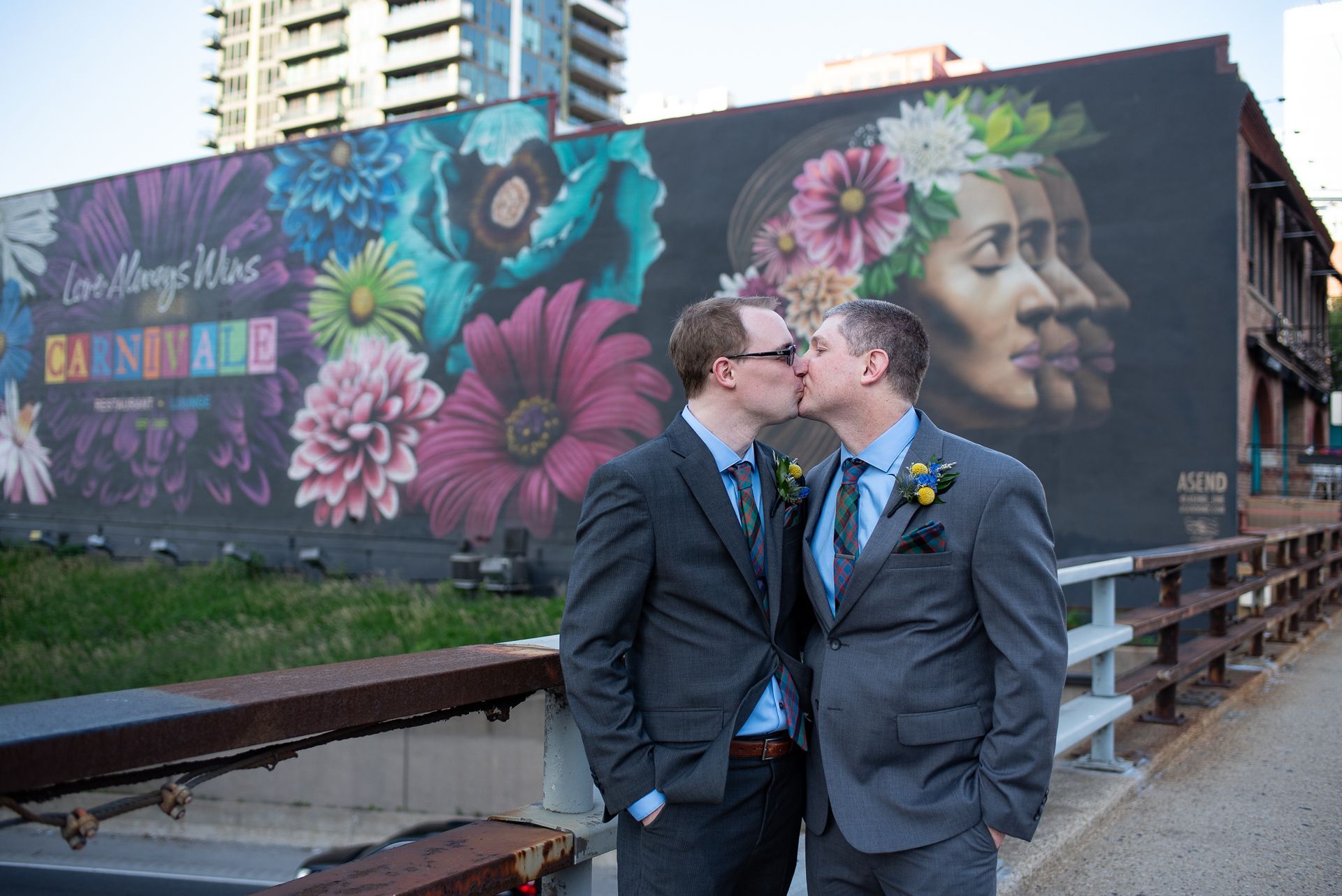 Two men in suits kissing in front of a colorful mural.