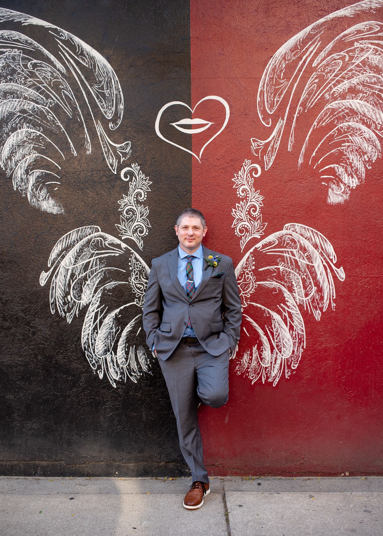 Man in grey suit leans against a wall with a painted rooster design, black and red backdrop.