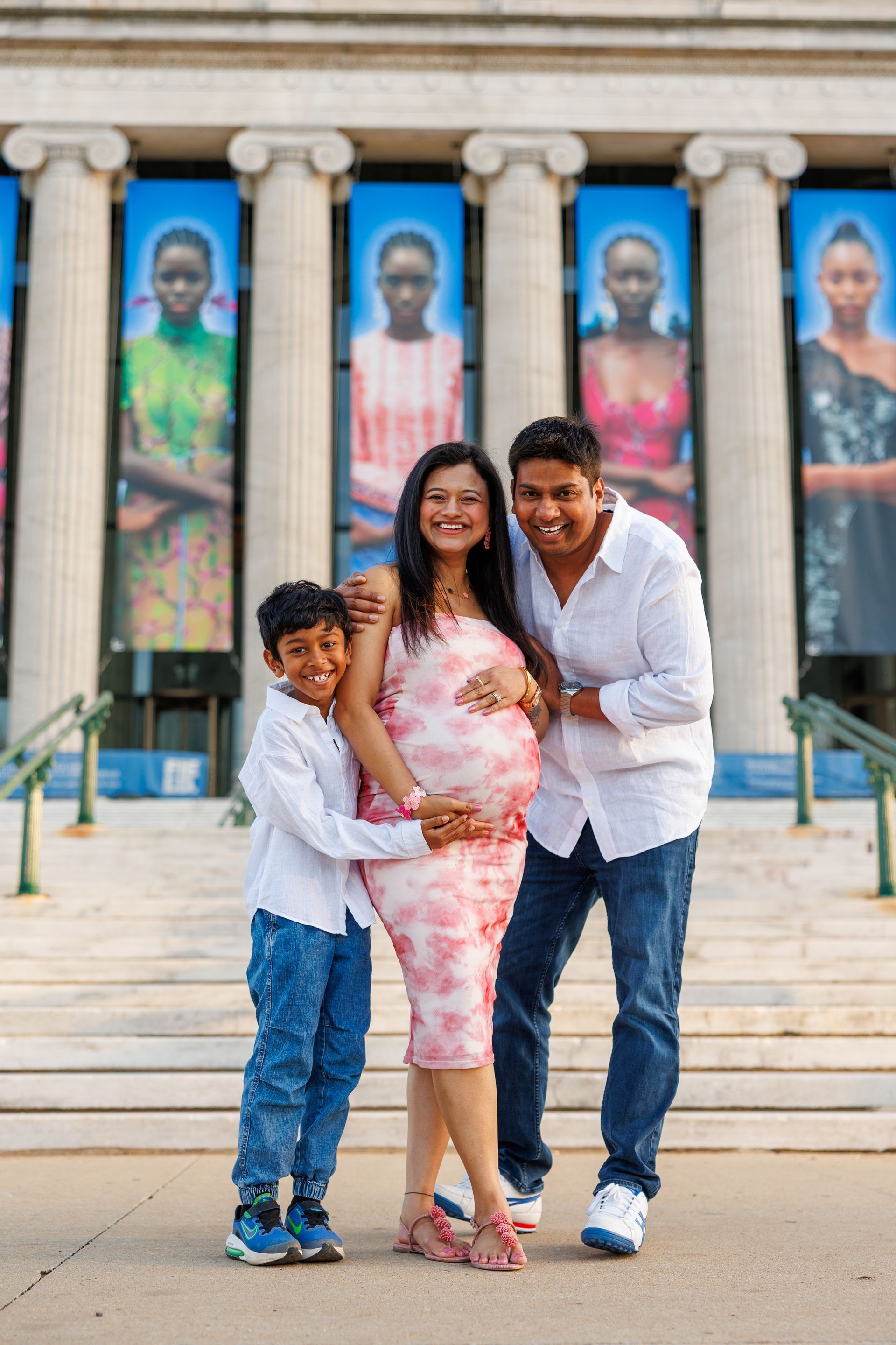 Family of three smiling, posing in front of a building with columns, pregnant woman holding belly.
