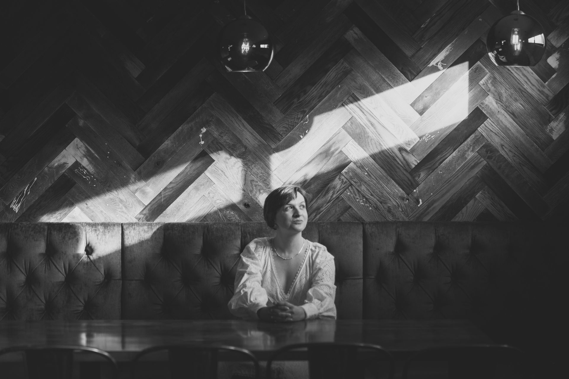 Woman seated at a table, looking up toward a beam of sunlight on a textured wall. Black and white.
