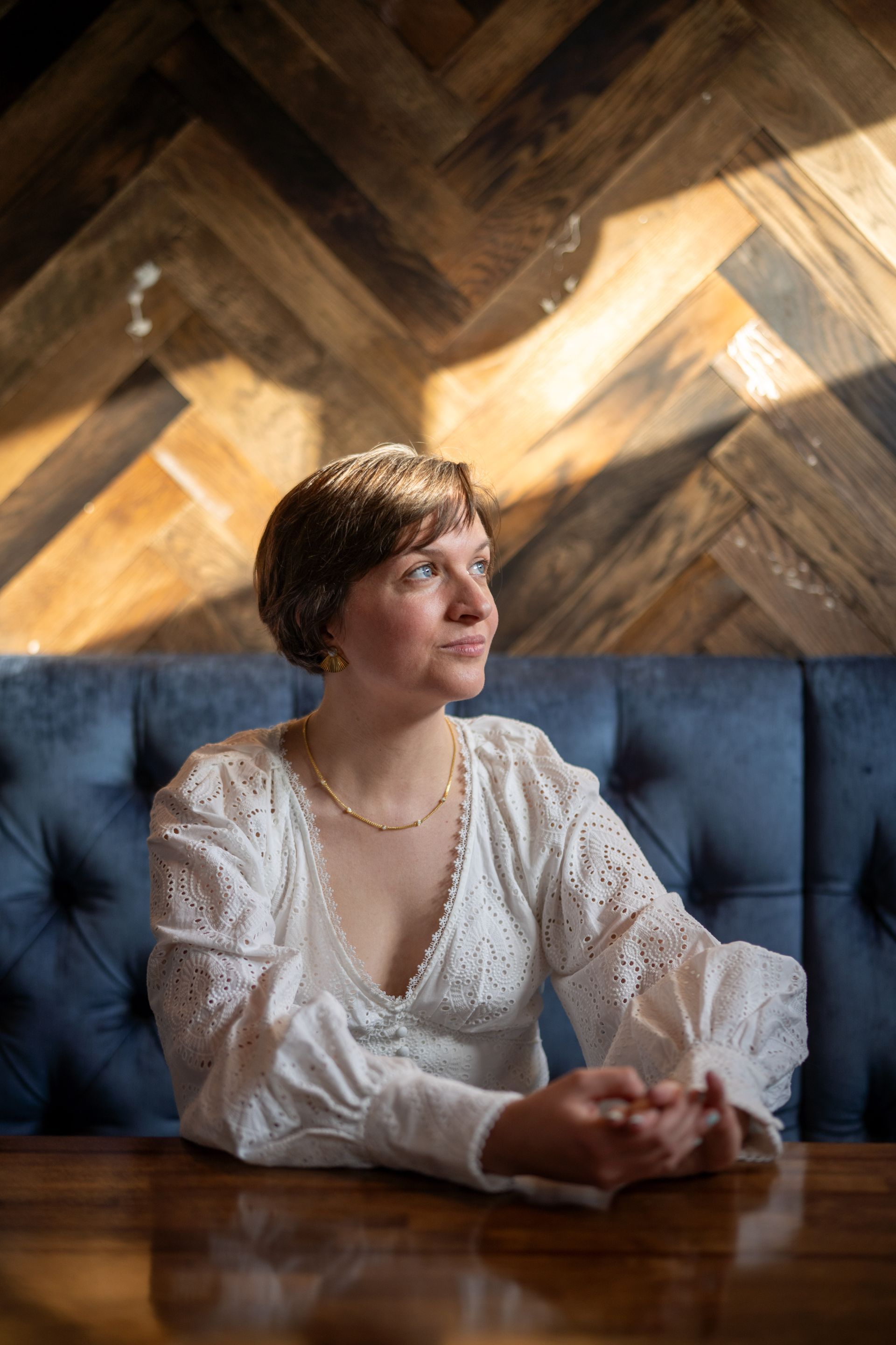Woman sitting at a table, looking off to the side, wearing a white blouse, in front of a blue booth and wood wall.