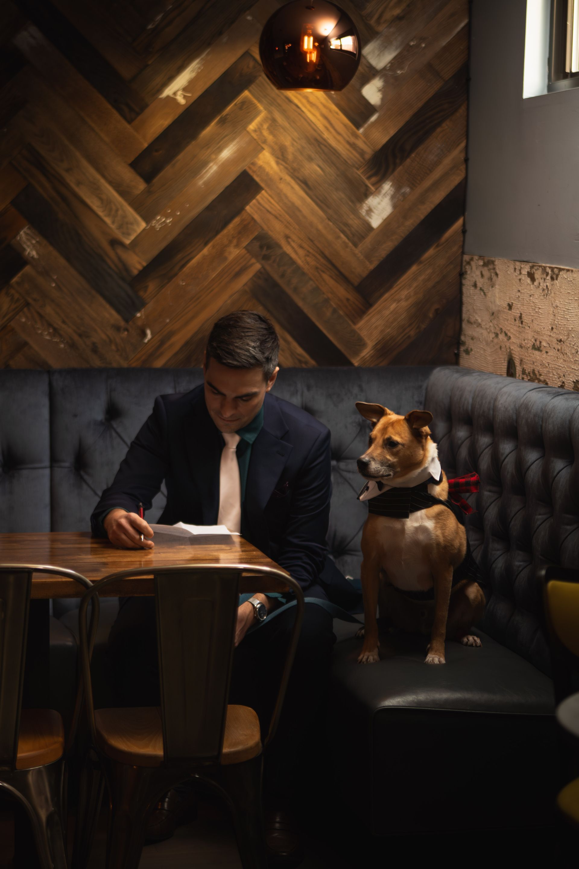 Man in suit reads at a table with a dog wearing a bandana, seated next to him in a dark booth.